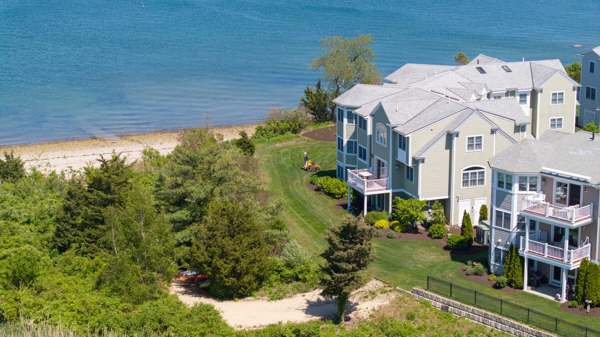 Coastal view with townhouses, lush garden, and a person on a lawnmower near the beach and vibrant blue water in the background.