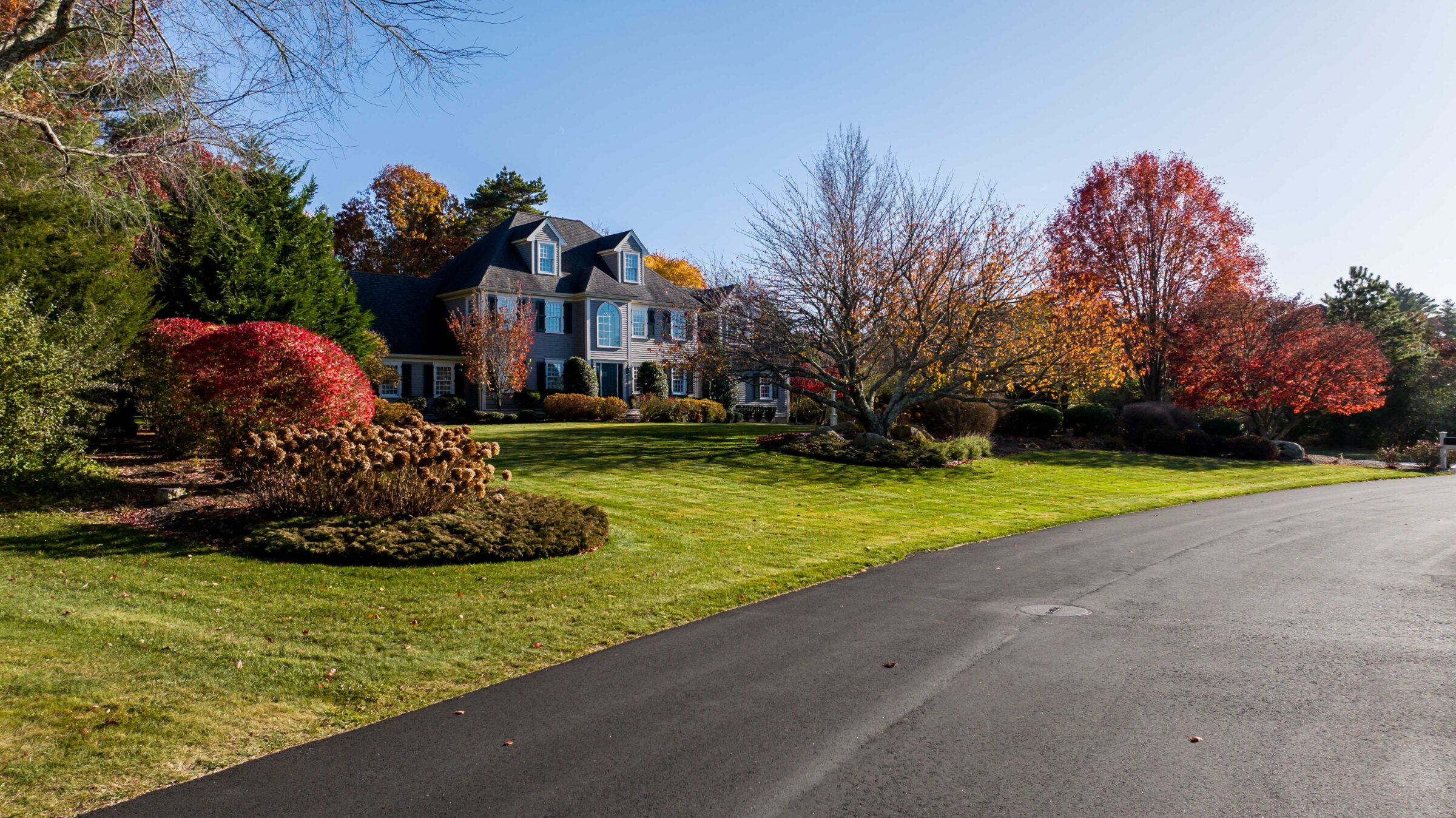Colonial-style house surrounded by colorful autumn trees, neatly manicured lawn, and a smooth asphalt road under clear blue sky.