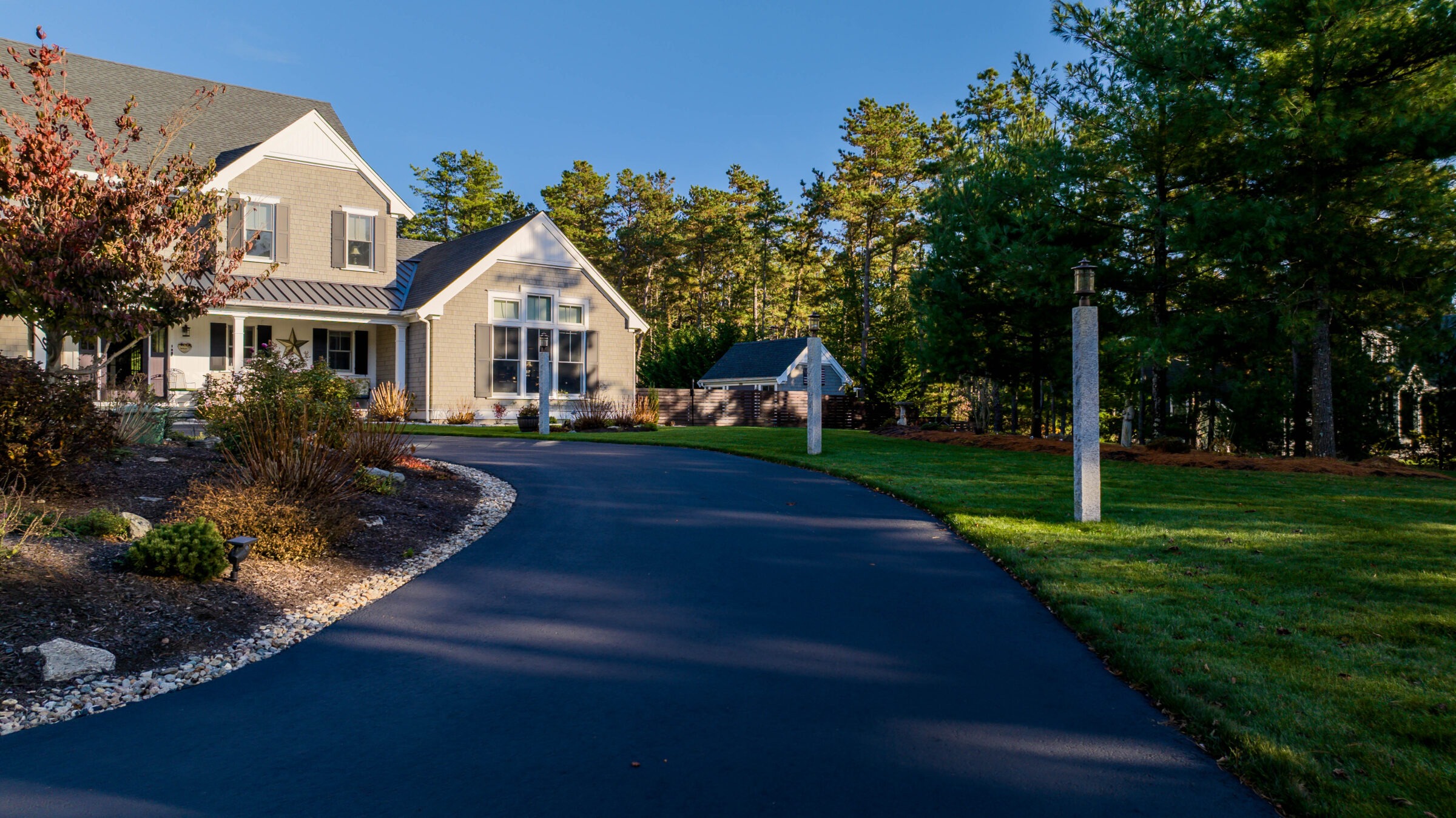 A charming house sits among trees, with a curved driveway leading to a two-story structure. Lush greenery and clear skies surround the scene.