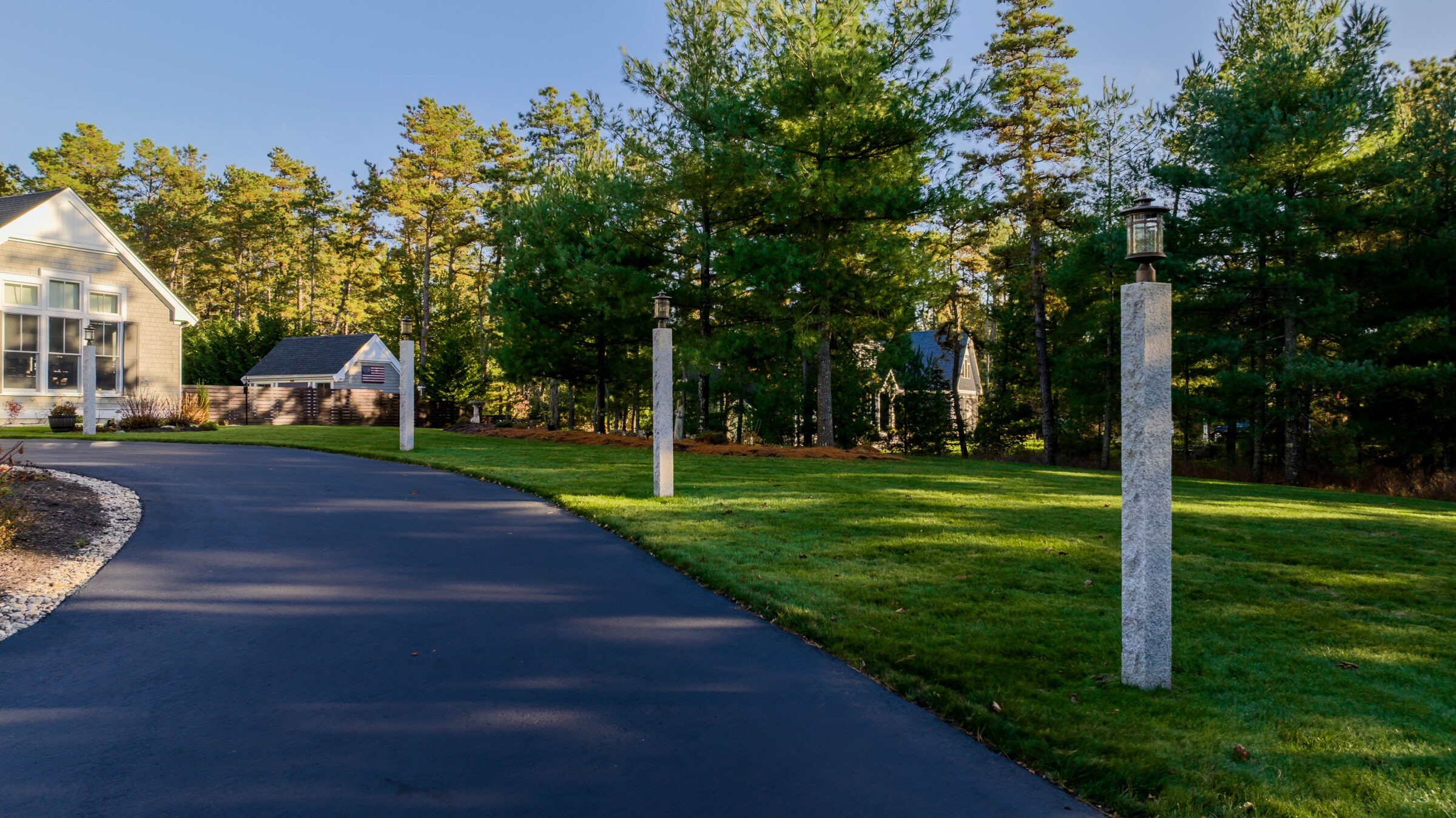 A suburban house with manicured lawn, surrounded by trees. A curved driveway with stone lampposts leads to the entrance.