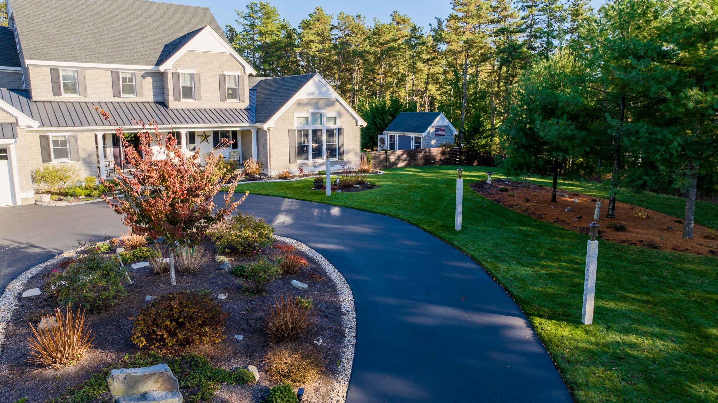 Suburban house with a manicured lawn and curved driveway, surrounded by trees and gardens, under a clear blue sky.