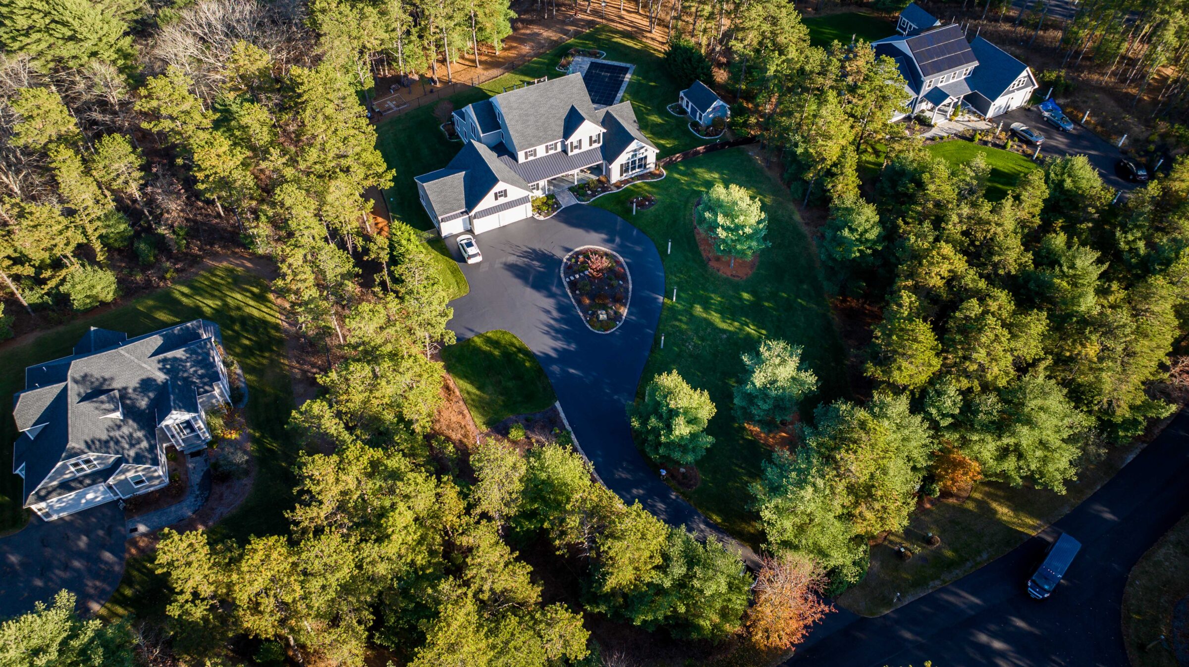 Aerial view of a suburban neighborhood with three houses, surrounded by dense trees, curving driveways, and a well-kept lawn.