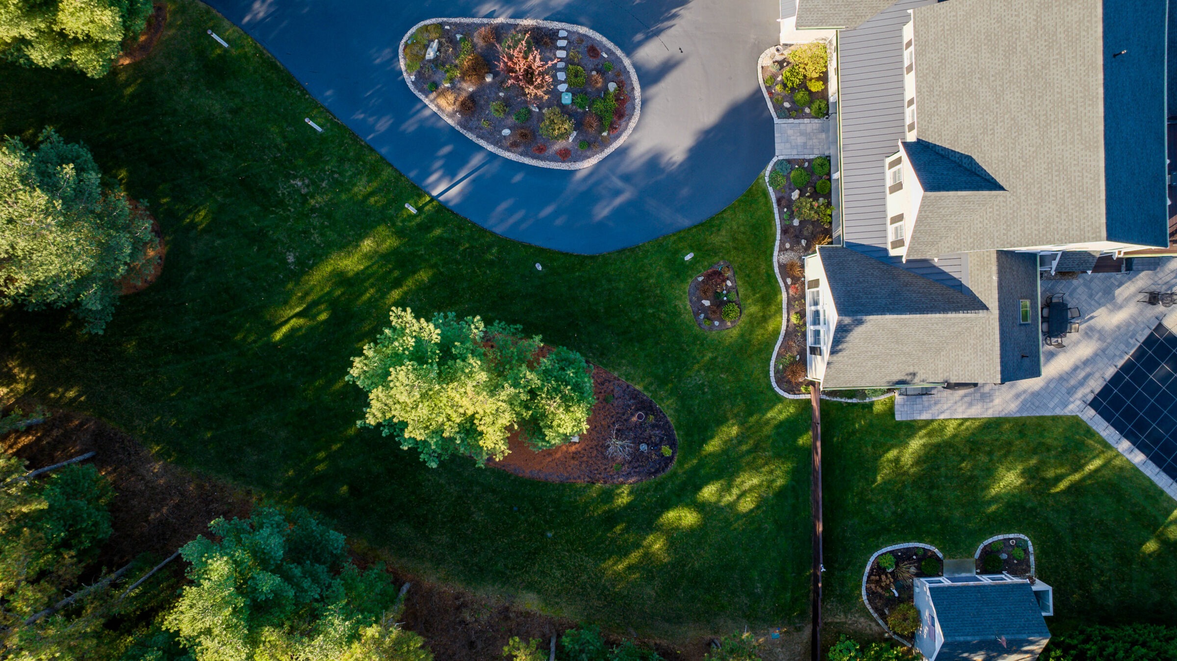 Aerial view of well-maintained modern house with landscaped garden, driveway, and diverse trees, highlighting symmetrical design and lush greenery under sunlight.