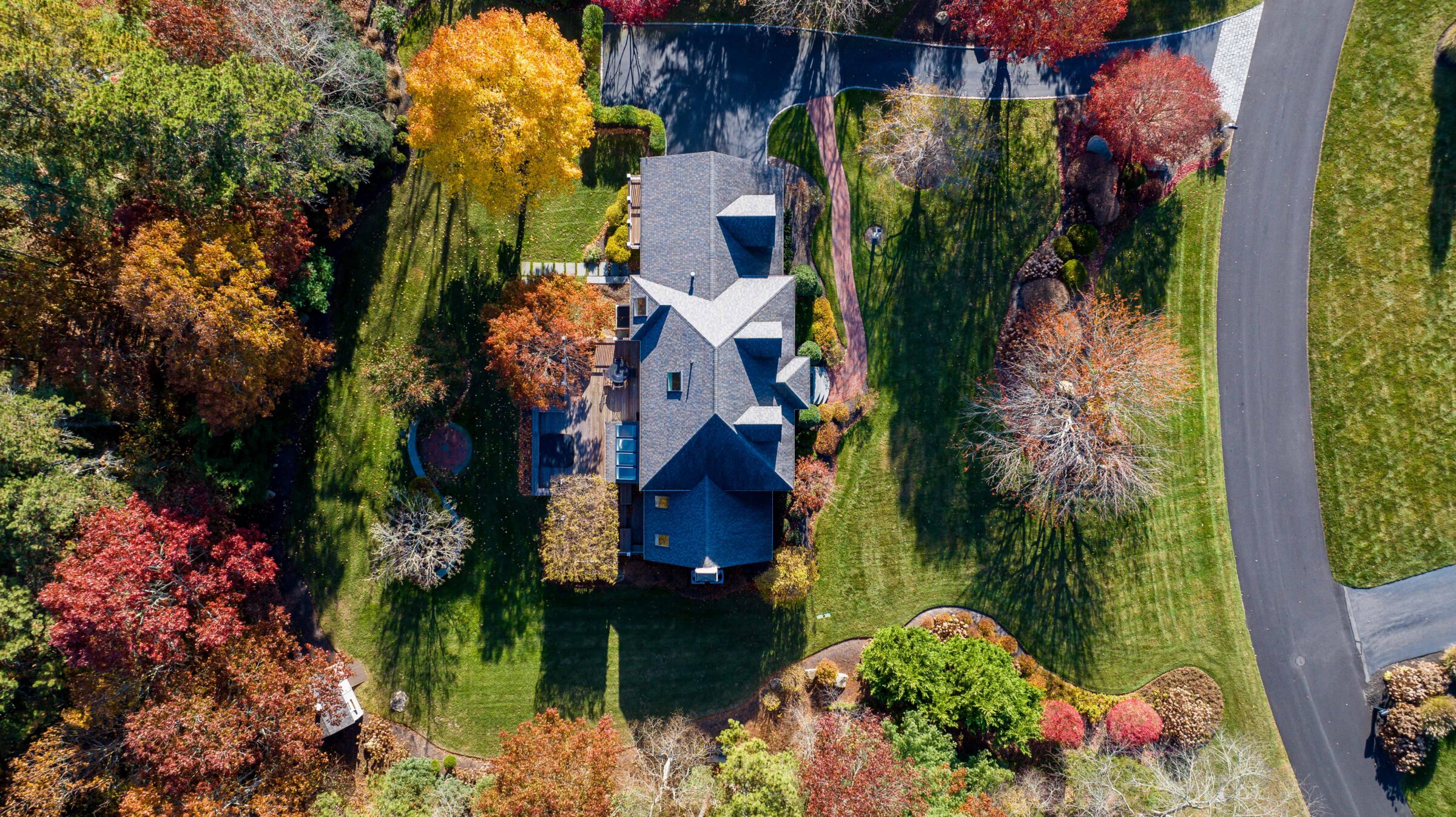 Aerial view of a large house surrounded by colorful autumn trees, lush garden, and a winding driveway on a sunny day.
