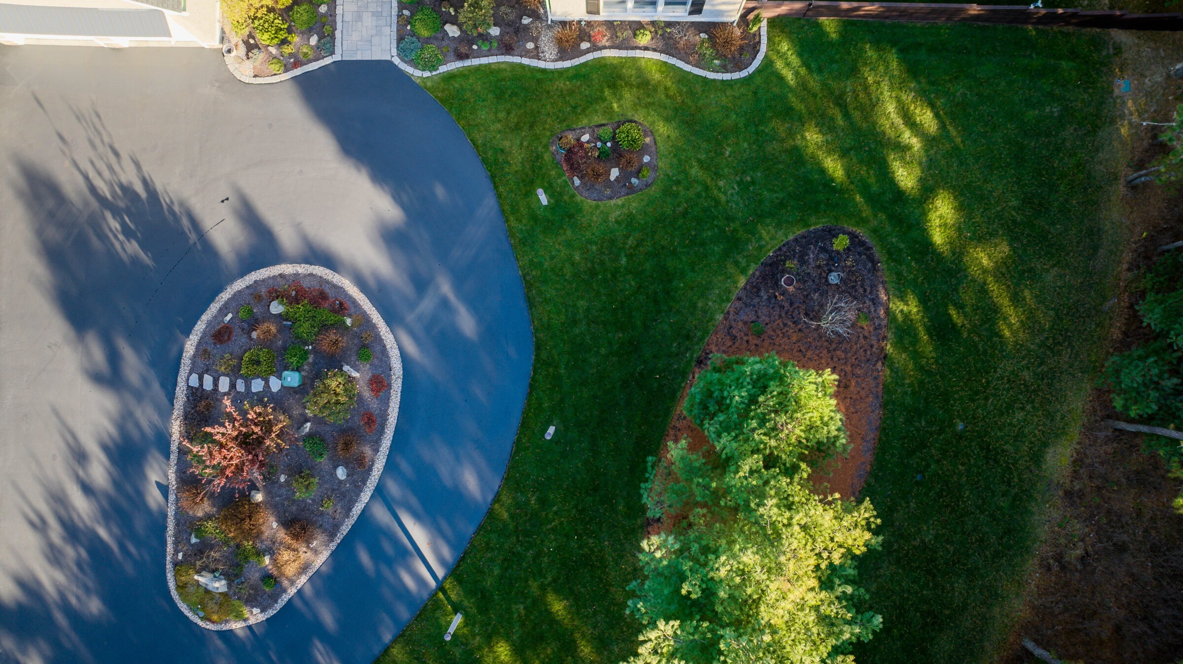 Aerial view of a residential driveway with curved paths, surrounded by landscaped greenery, trees, and shrubs on a sunny day.