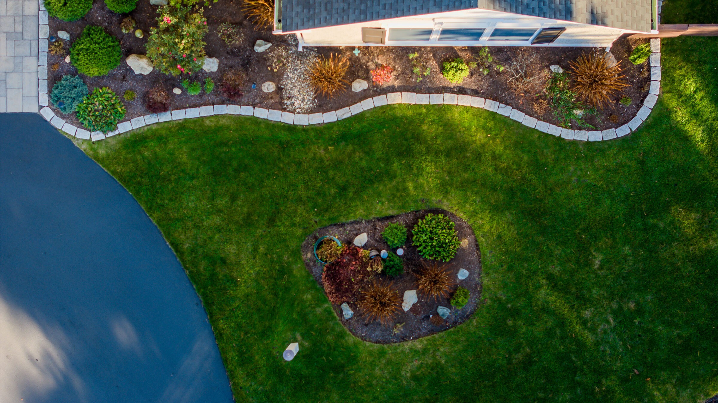 Aerial view of a landscaped garden featuring green grass, shrubs, and a stone-bordered flower bed, adjacent to a paved surface.
