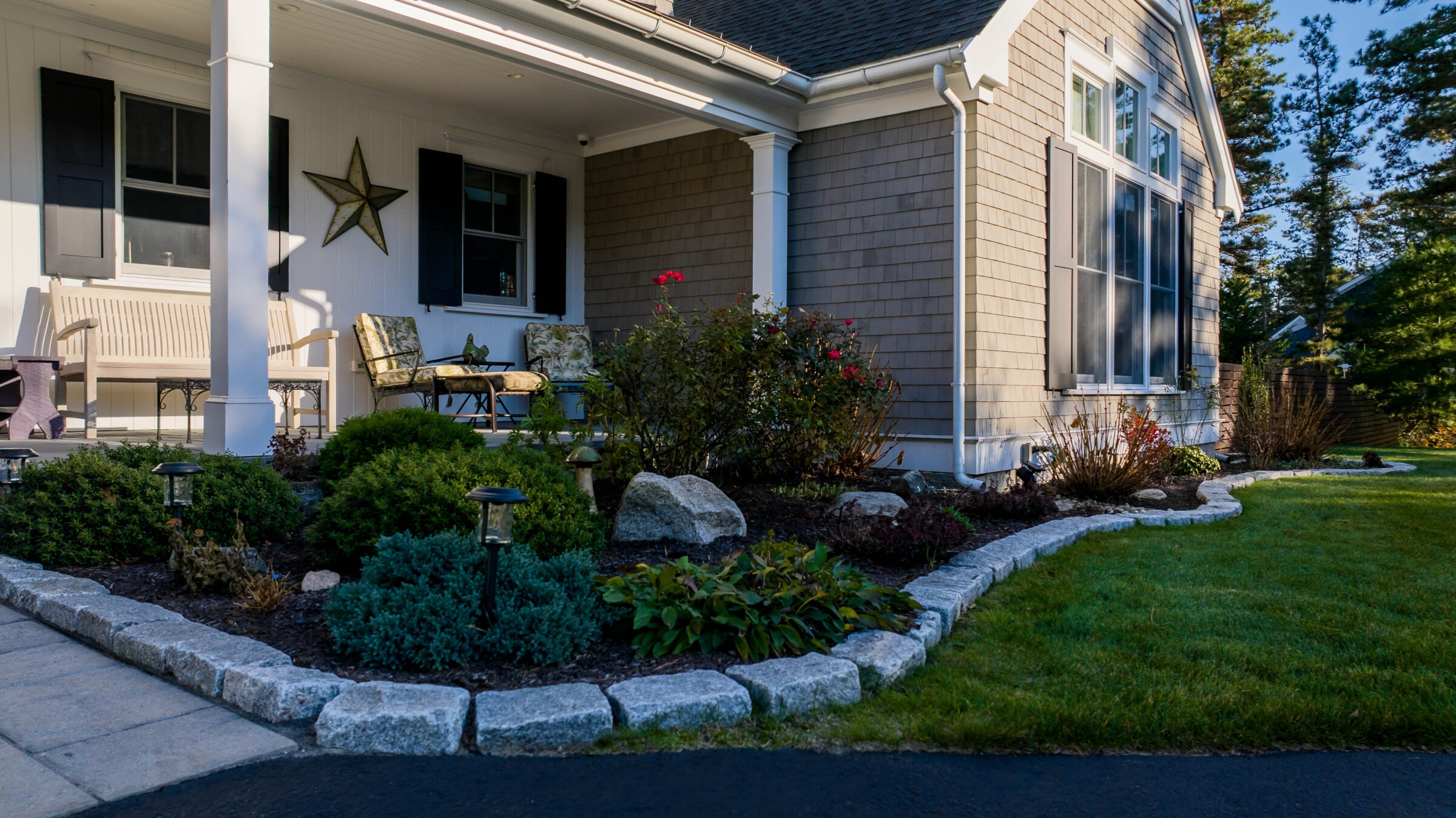 Cozy house porch with chairs and star decoration, surrounded by landscaped garden featuring green bushes and stone borders under a clear blue sky.
