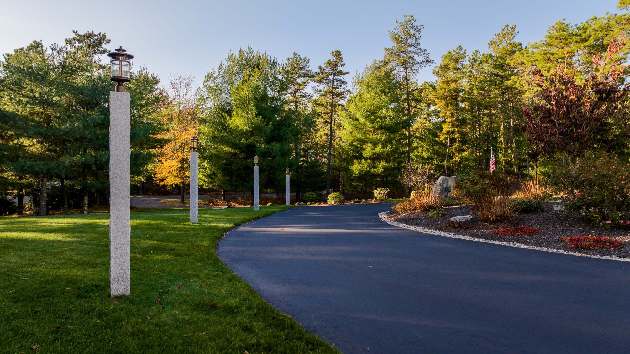 A paved driveway curves between green lawns and tall trees, bordered by tall lamp posts and a well-maintained garden under a clear sky.