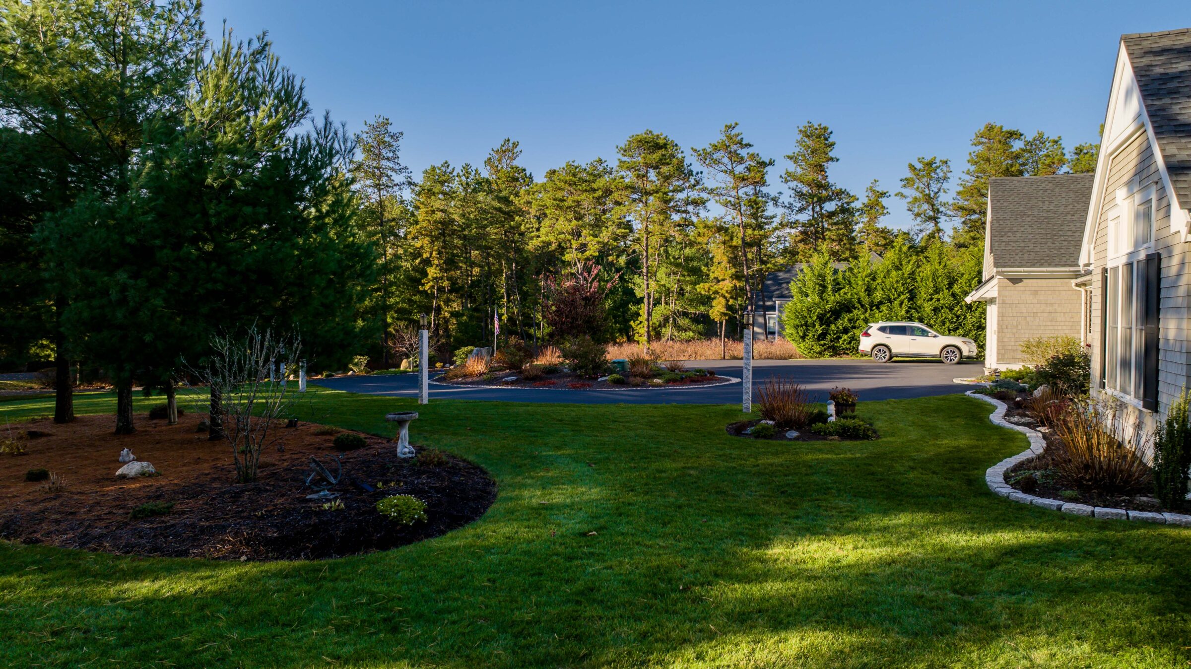 A serene suburban scene shows a neatly landscaped yard, trees, a parked car, and a residential house under a clear blue sky.