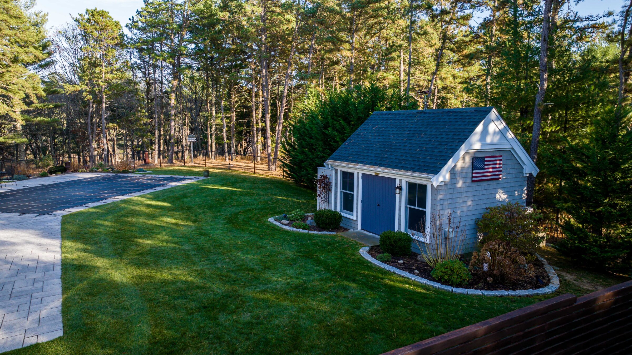 A small shed with an American flag stands in a backyard, next to a covered pool, surrounded by trees and grass.