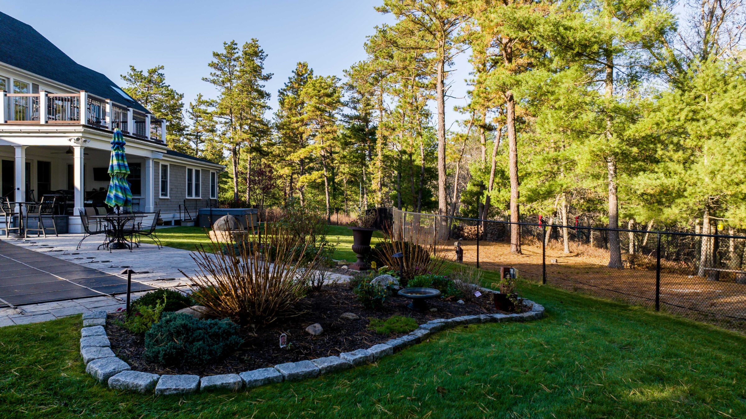 A backyard garden with a patio, outdoor furniture, and wooded area. No people or recognizable landmarks are visible.