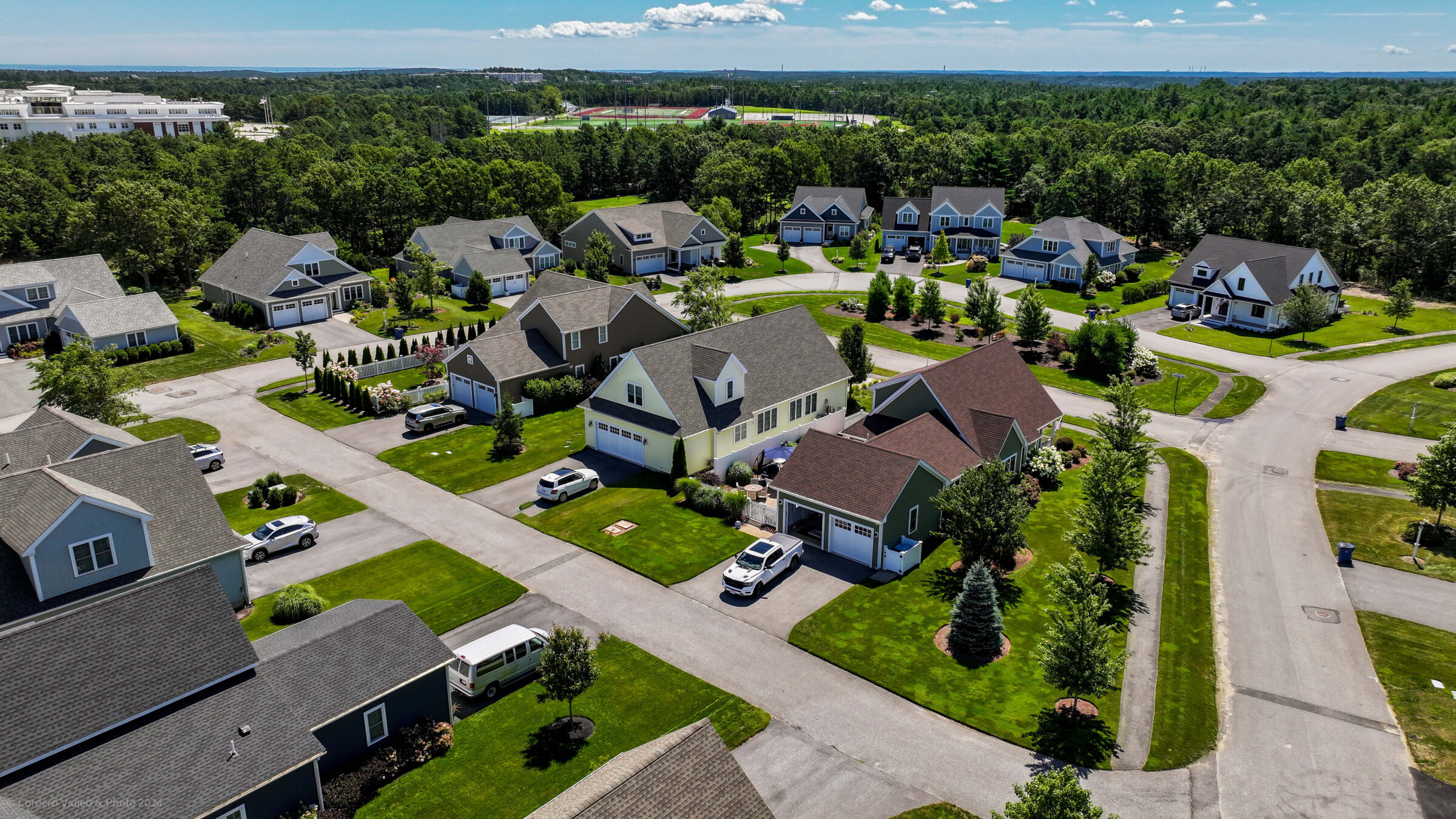 Aerial view of a suburban neighborhood with well-maintained houses, green lawns, and parked cars, surrounded by trees and a distant sports field.