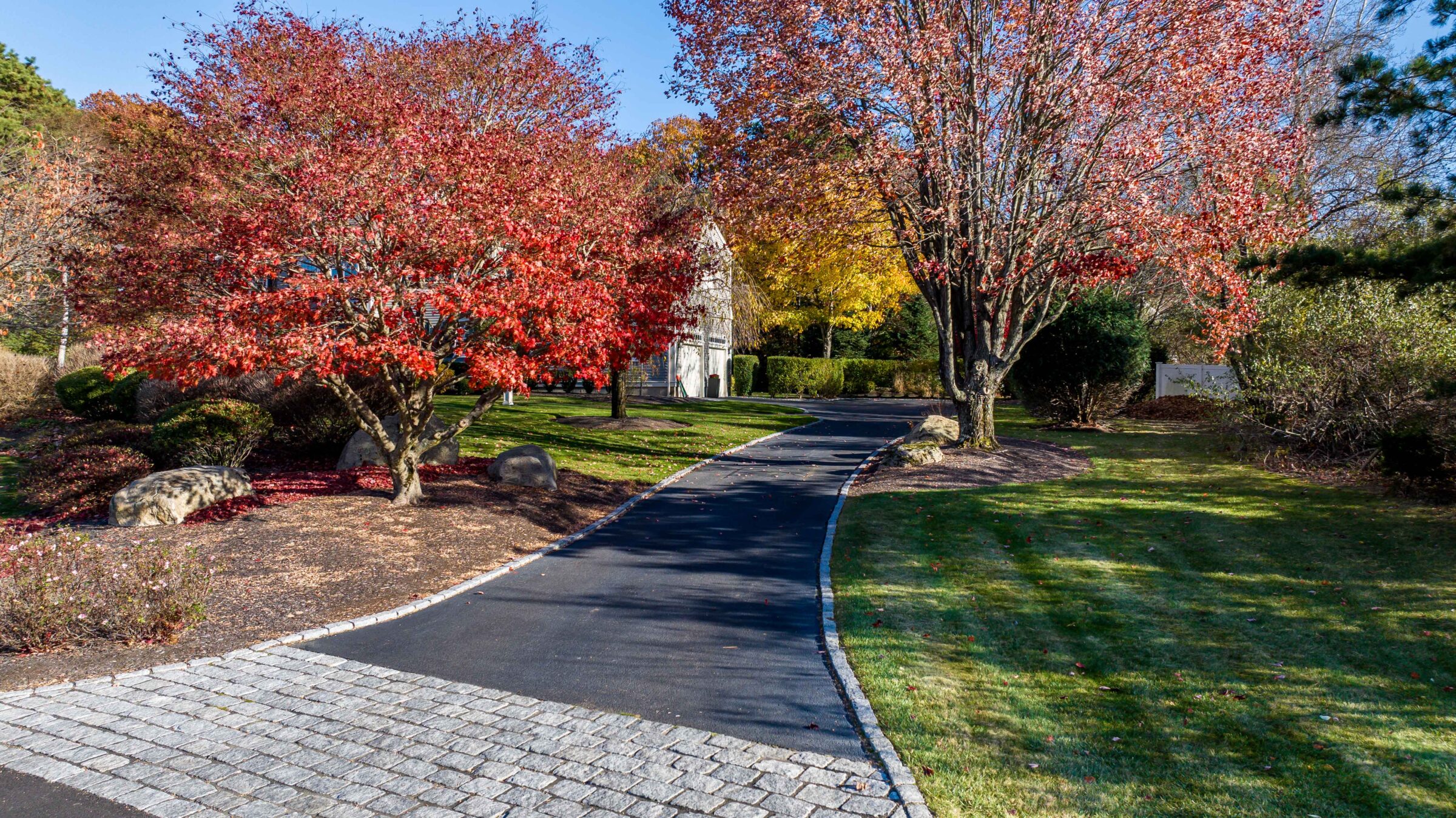 A paved path curves through a park with vibrant autumn trees. A small stone building is seen in the distance under clear skies.