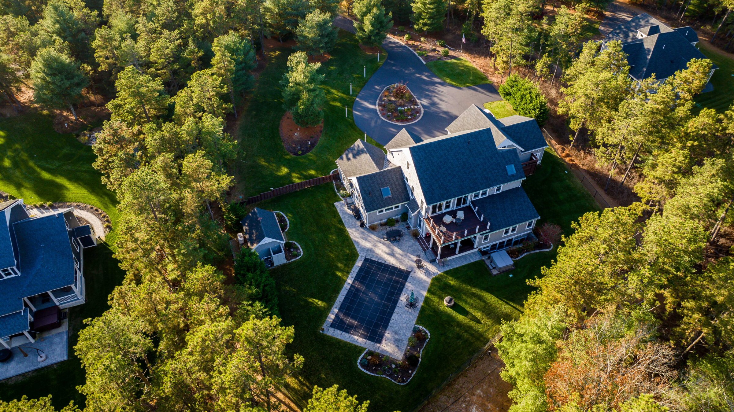 Aerial view of a large house with a pool in a forested area, surrounded by trees, and a winding driveway.