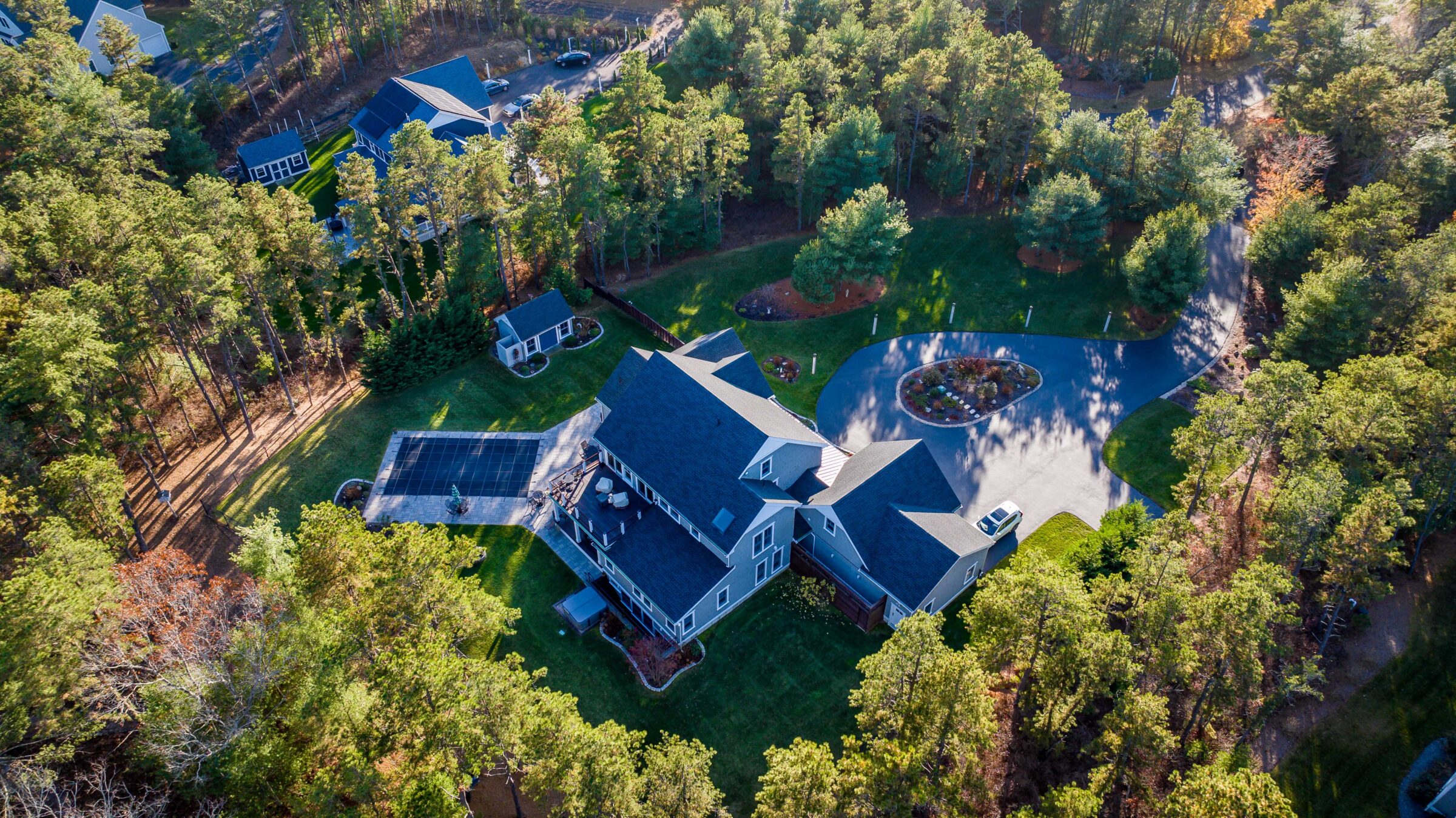Aerial view of a large house surrounded by dense trees, a driveway, and landscaped gardens in a serene forested area.