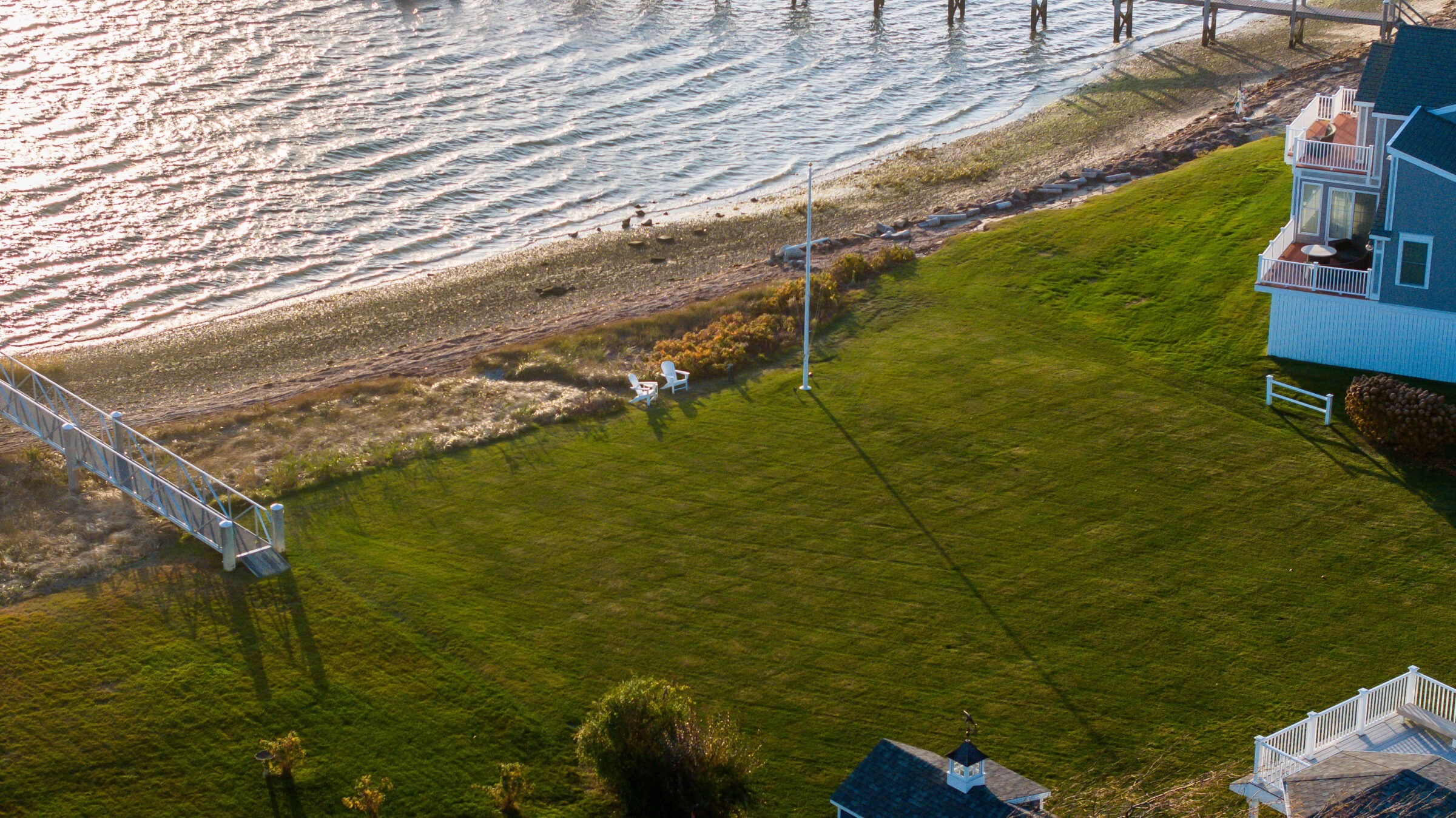 Aerial view of a coastal property with a lawn, chairs, small pier, and adjacent house by a serene, sunlit shoreline.