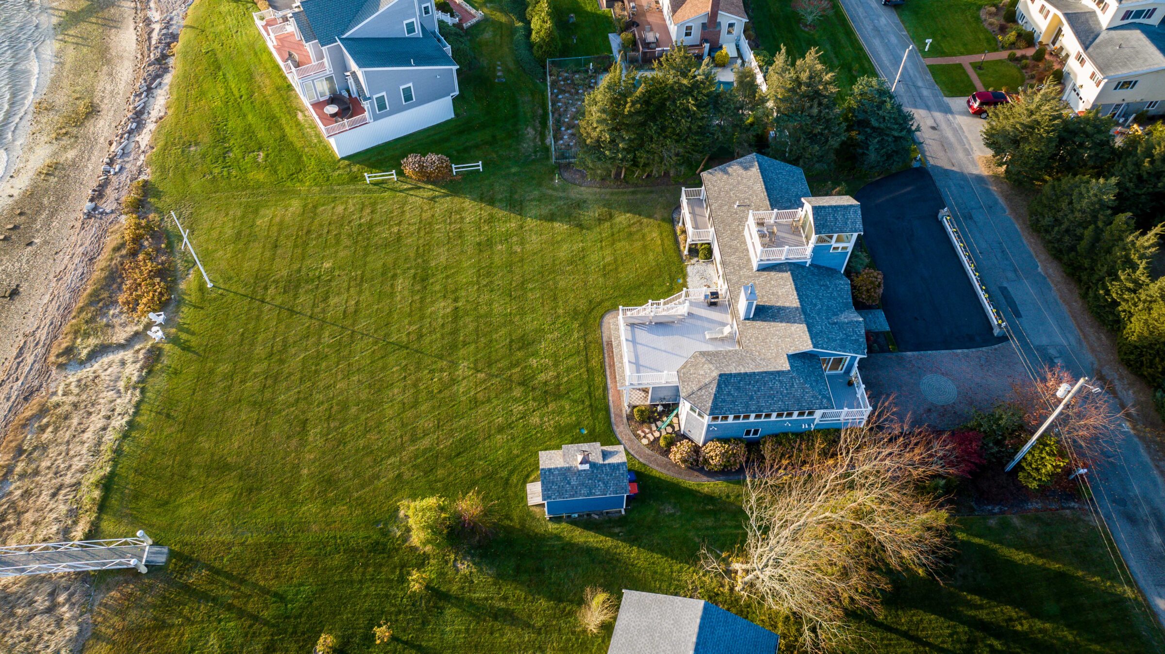 Aerial view of coastal residential area with large green lawn, houses, and a beach. Trees and roads surround the homes, creating a serene atmosphere.