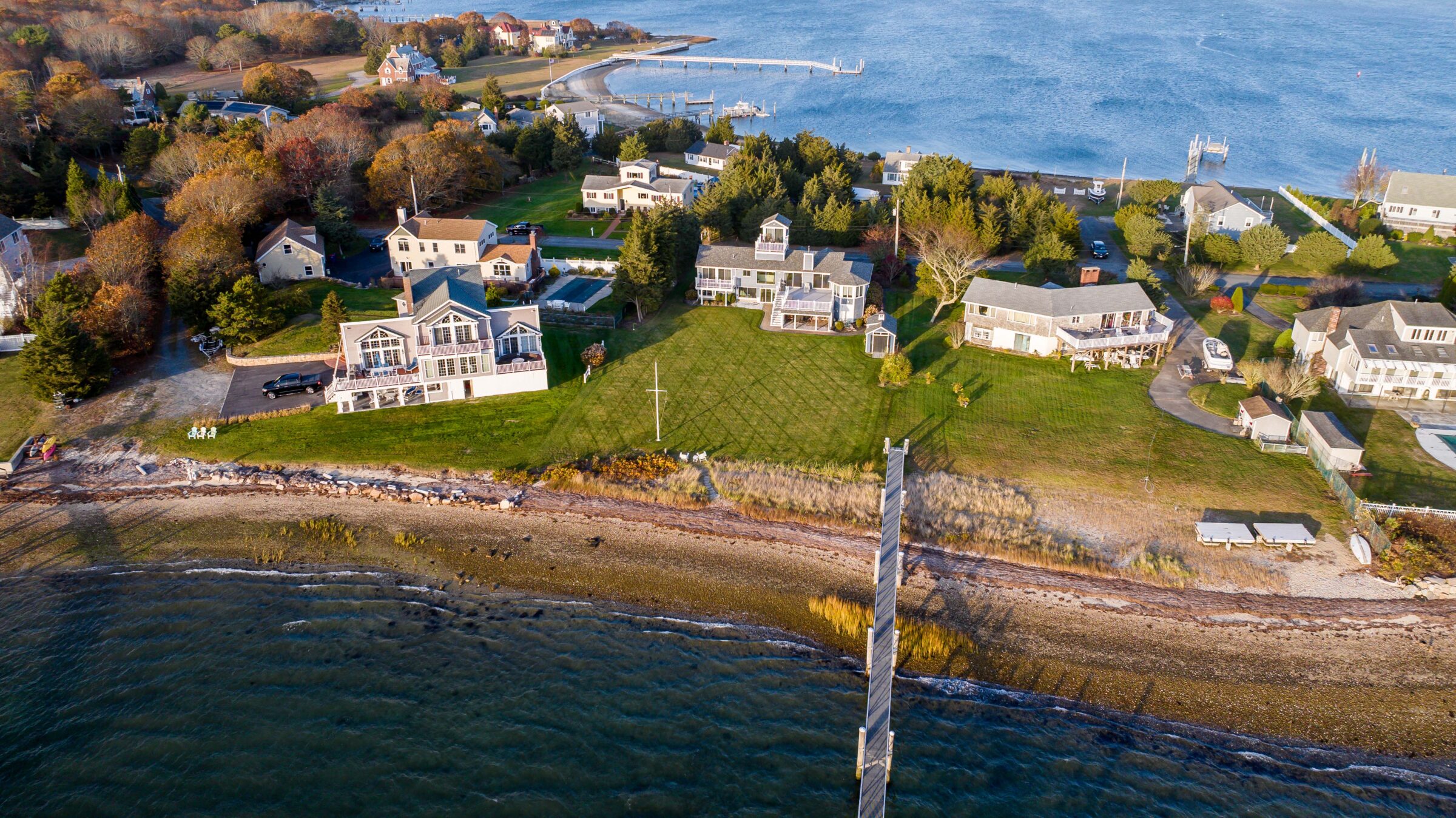 Aerial view of a coastal neighborhood with houses, green lawns, and a waterfront dock. No recognizable landmarks or historical buildings visible.