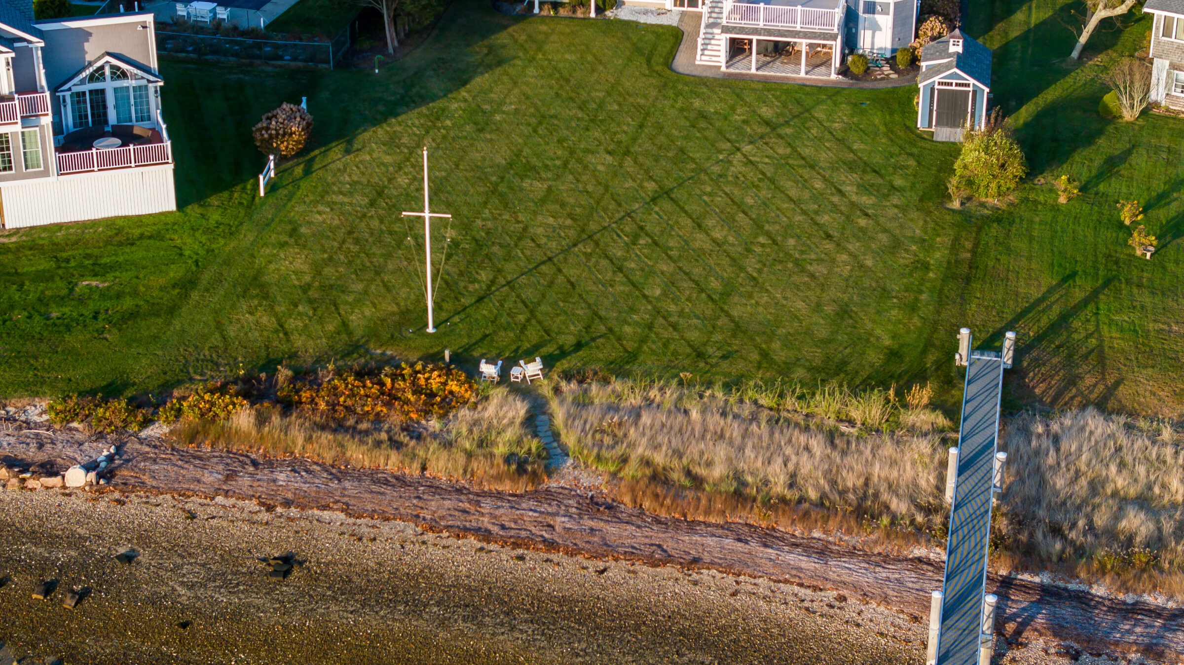 Coastal green lawn with chairs, a flagpole, and houses. Wooden pier extends into a rocky shoreline. No people visible.