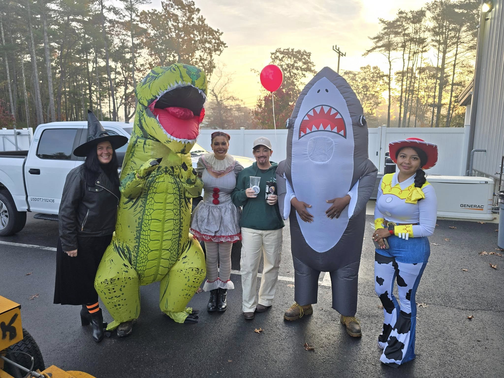 A group of people in costumes, including a dinosaur and a shark, standing outdoors near a truck, surrounded by trees.