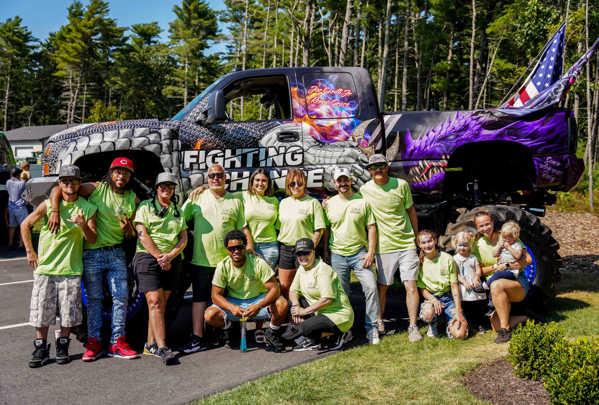 A group of people in matching green shirts stand in front of a large, colorful truck with a dragon design in a park setting.