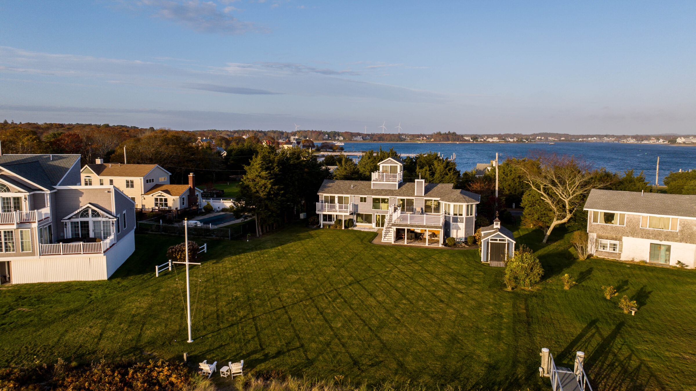 Coastal houses with spacious lawns overlook calm waters and distant shoreline. Sparse trees surround the area under a clear blue sky.
