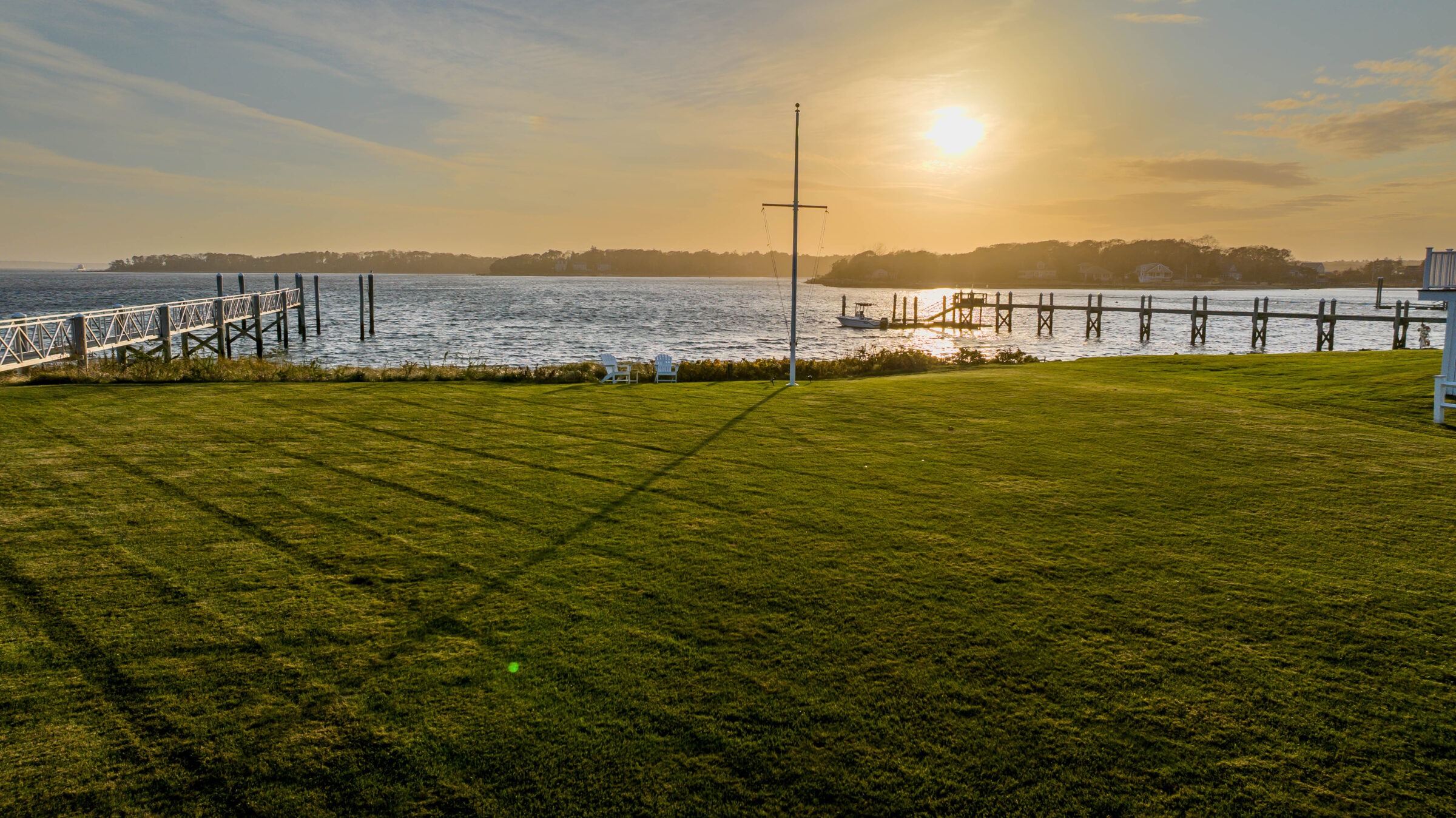 A serene coastal scene at sunset with a grassy lawn, wooden docks, and calm water. Two white chairs face the sunset.