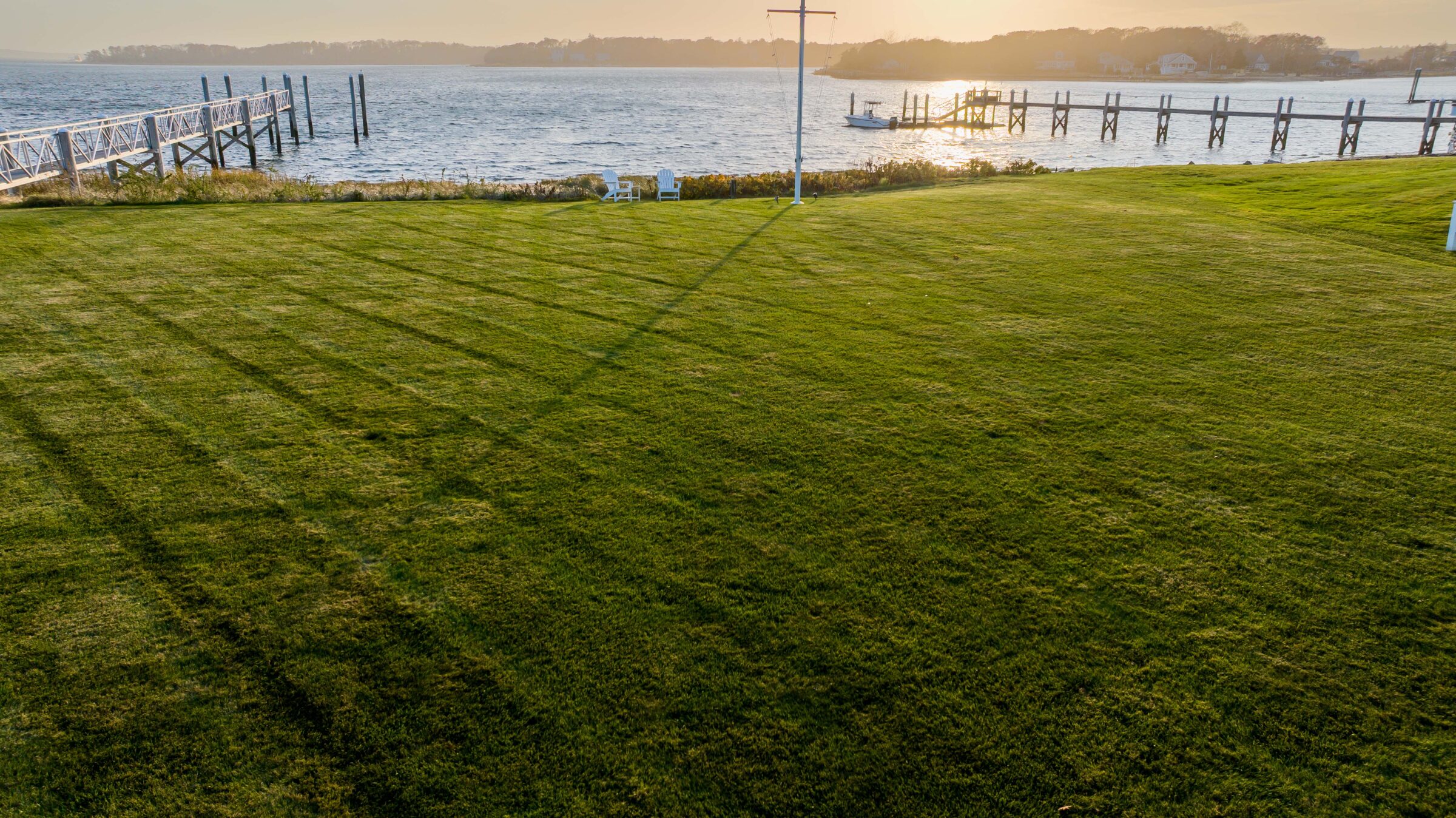 Lush green lawn with waterfront view, small dock, and Adirondack chairs facing the calm water under a clear sky at sunset.