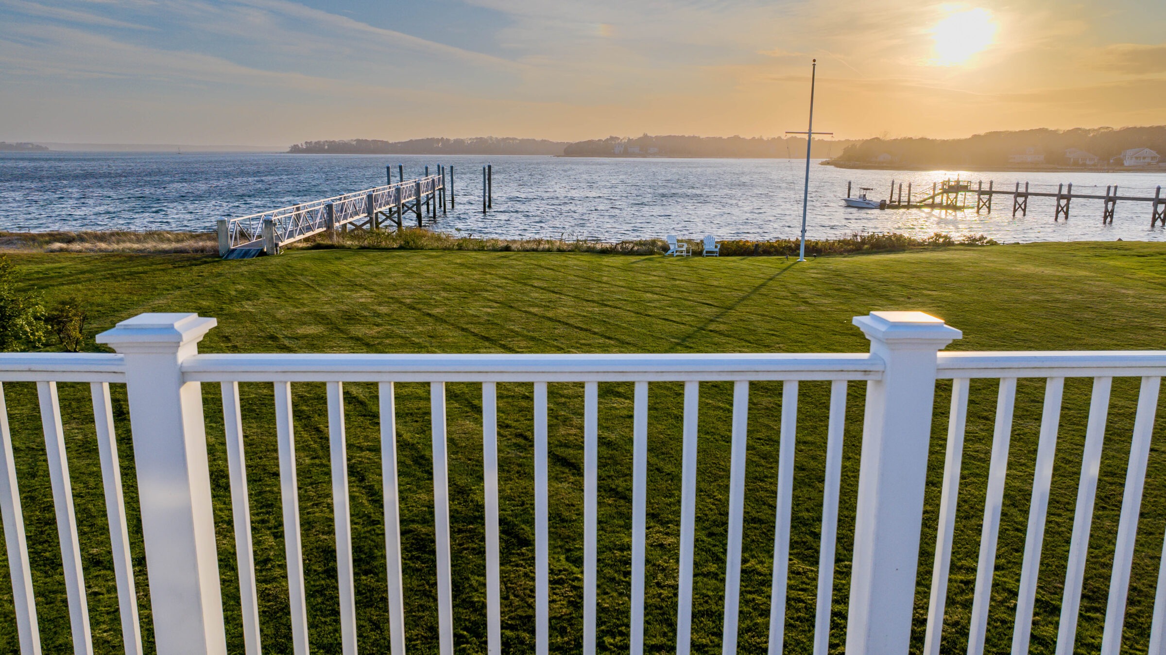 White fence overlooks a grassy yard and dock by the water, with sunset illuminating a peaceful, serene landscape in the background.