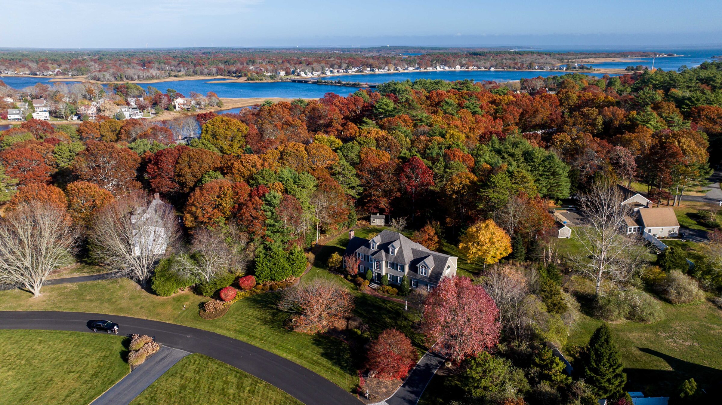 Aerial view of a suburban area with vibrant autumn foliage, houses, a winding road, and a person near a car by a lake.