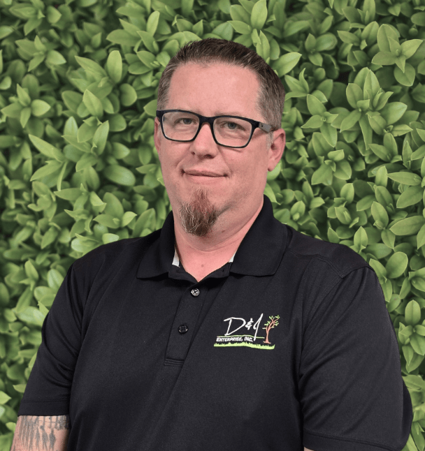 A person wearing glasses and a logoed polo shirt stands before a vibrant leafy background, smiling with arms crossed.