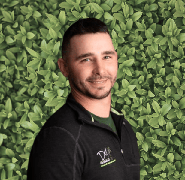 A person in a black jacket stands against a lush, leafy green background, smiling at the camera.