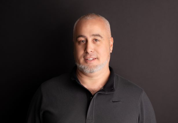 a person smiles against a black background. They have a clean white beard and a black golf shirt.