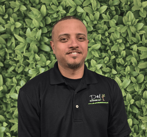 A person wearing a black polo shirt stands in front of a lush green leaf backdrop, smiling at the camera.