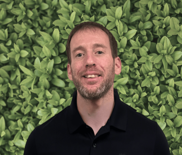 A person with a beard and short hair smiles in front of a leafy green background, wearing a black shirt.