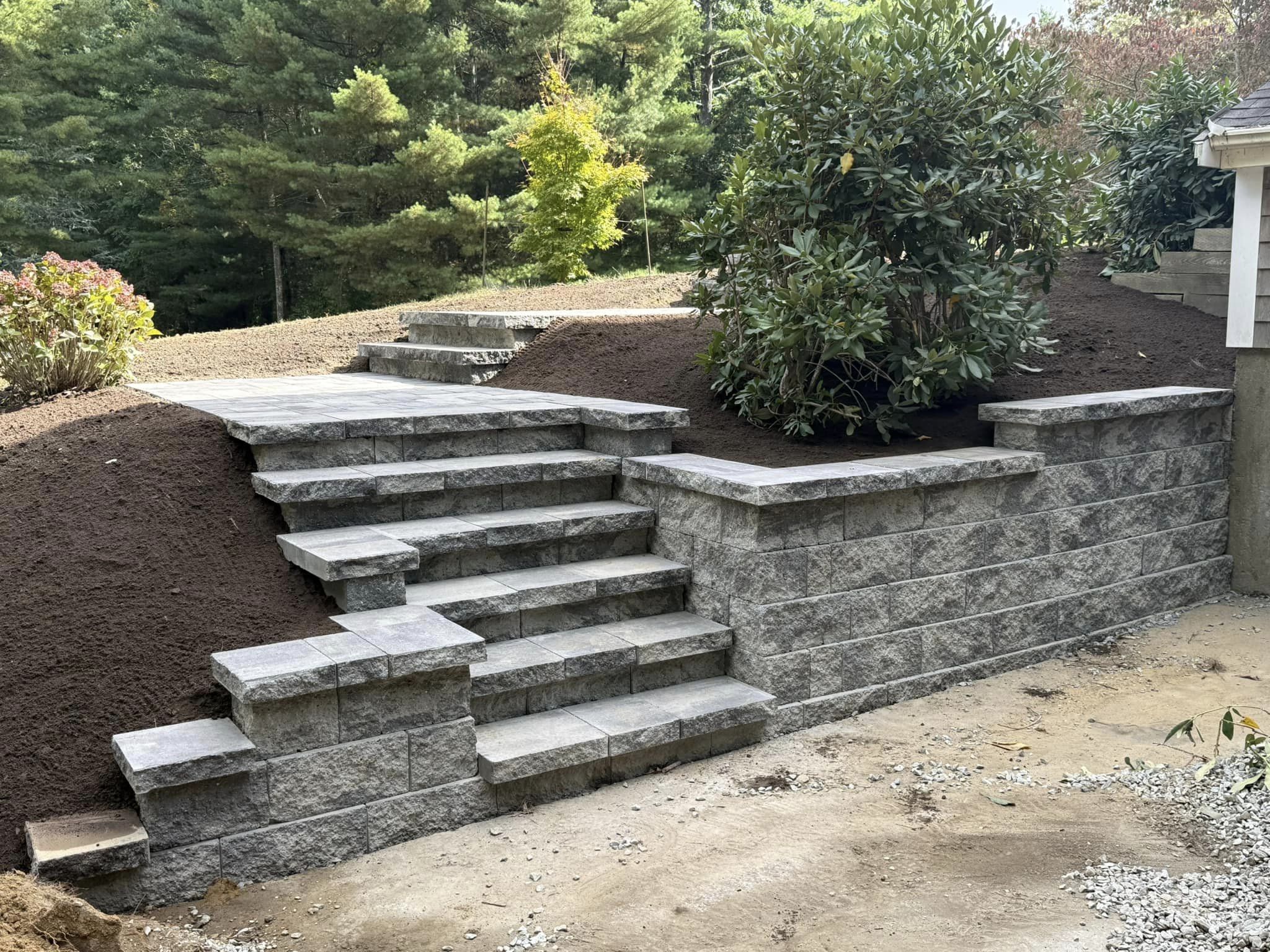Stone steps ascend through landscaped earth, bordered by shrubs, leading to a wooded area with lush green trees in the background.