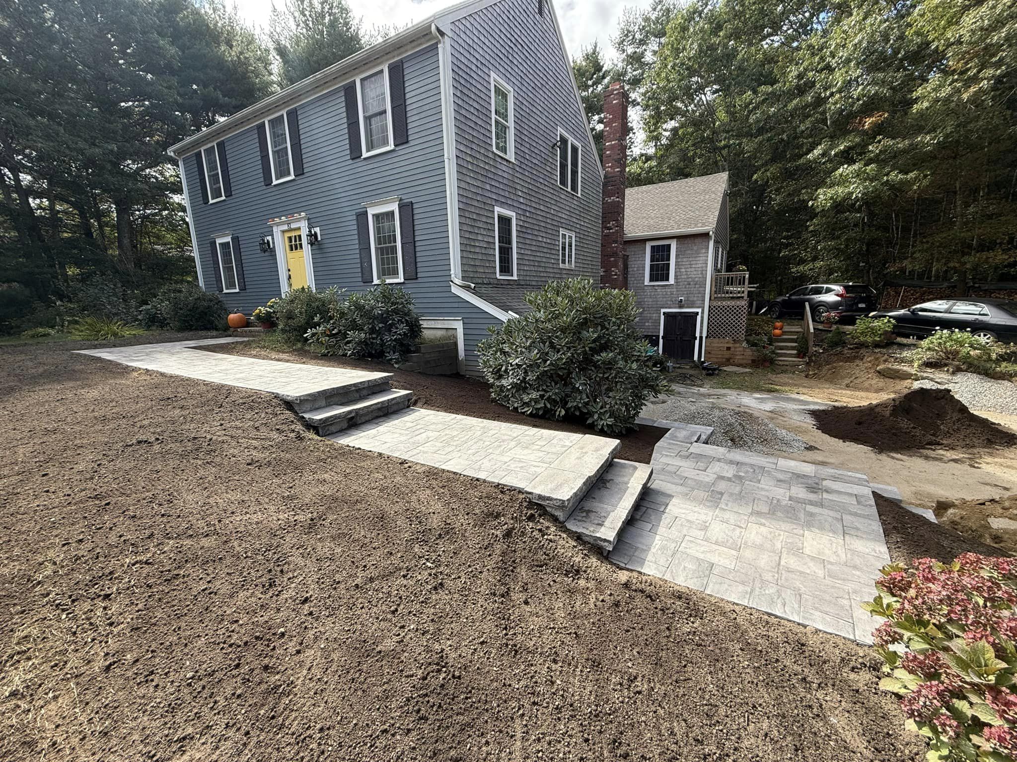 A blue house with a yellow door, surrounded by trees, features a new gray stone walkway and steps, near parked cars and landscaping.