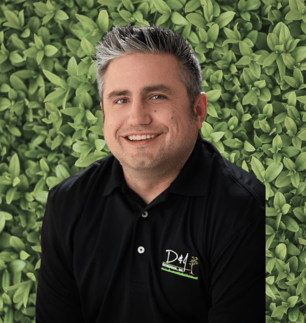 A person in a black polo shirt smiles against a leafy green background, displaying a logo with a tree motif on the chest.