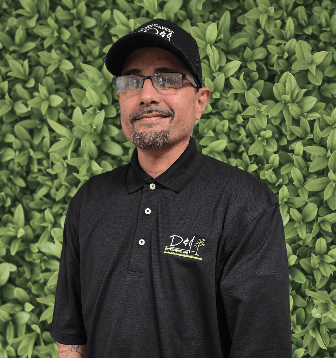 A person in a black polo and cap stands smiling in front of a leafy green wall, with embroidered logos visible.