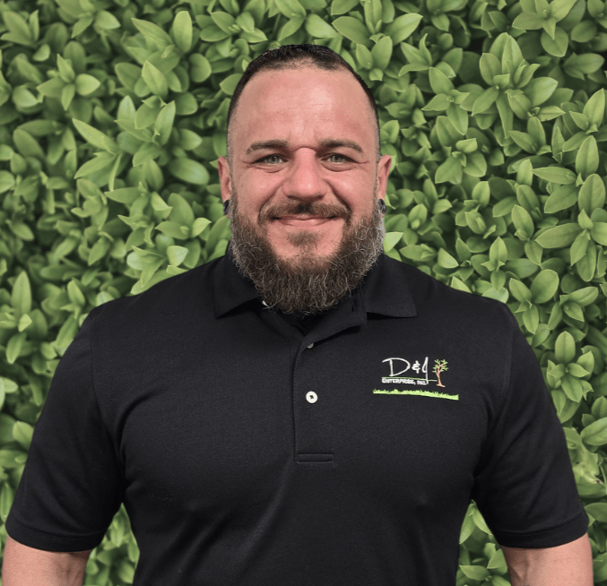 A person with a beard, wearing a black polo shirt, stands smiling in front of a leafy green background.