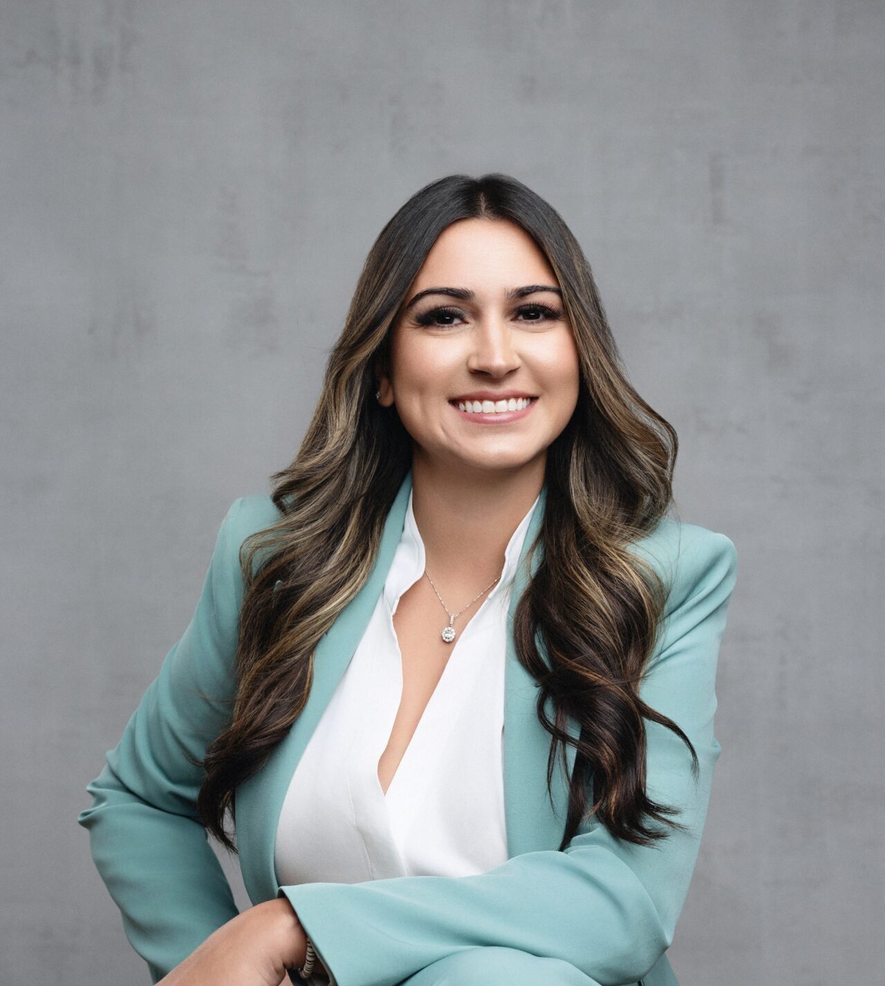 A person in a light teal suit smiles against a plain gray background, showcasing long wavy hair and a minimalist necklace.