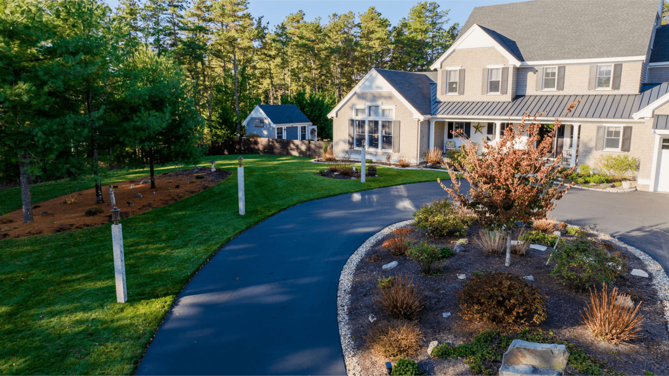 a house with a beautiful landscaped lawn as well as a garden in the center of a driveway roundabout featuring shrubs, a tree, and a stone trim.