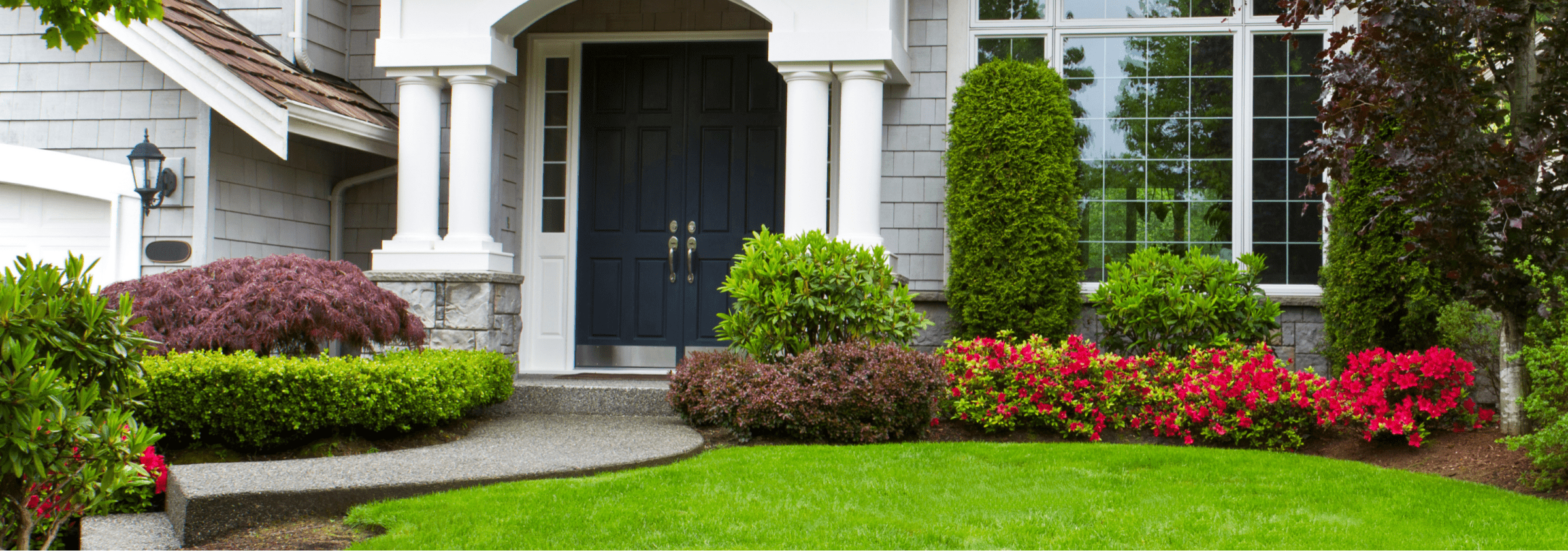 Modern home entrance with double doors, surrounded by manicured greenery and colorful flowerbeds. Symmetrical pillars enhance the welcoming, elegant facade.