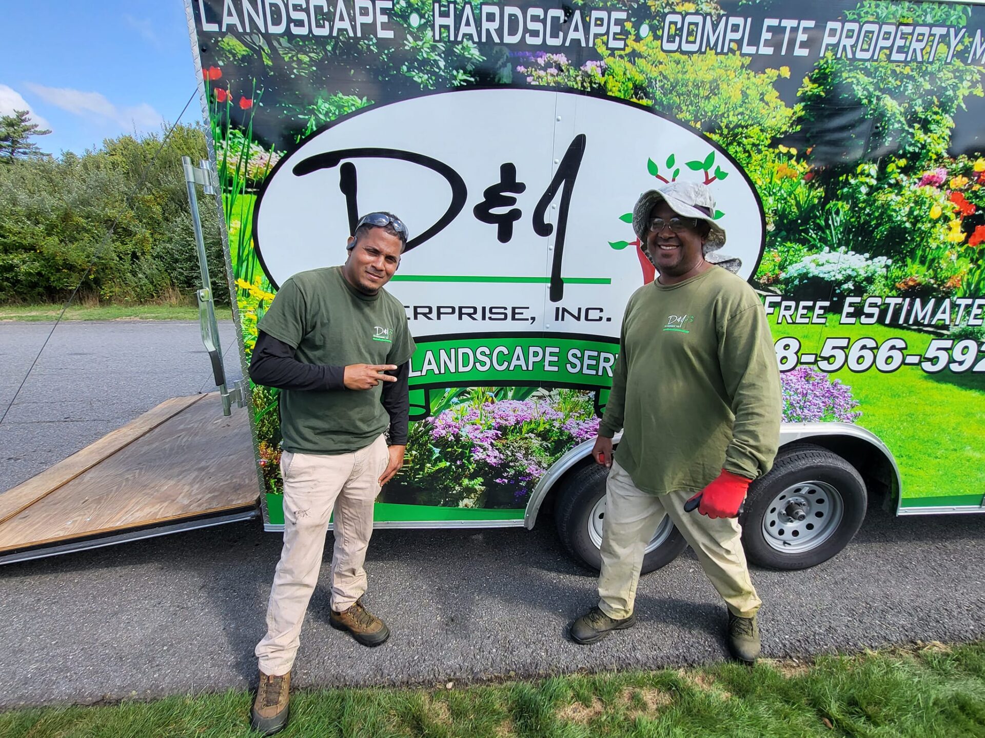 Two people in matching green outfits stand smiling in front of a colorful landscaping service trailer on a sunny day.