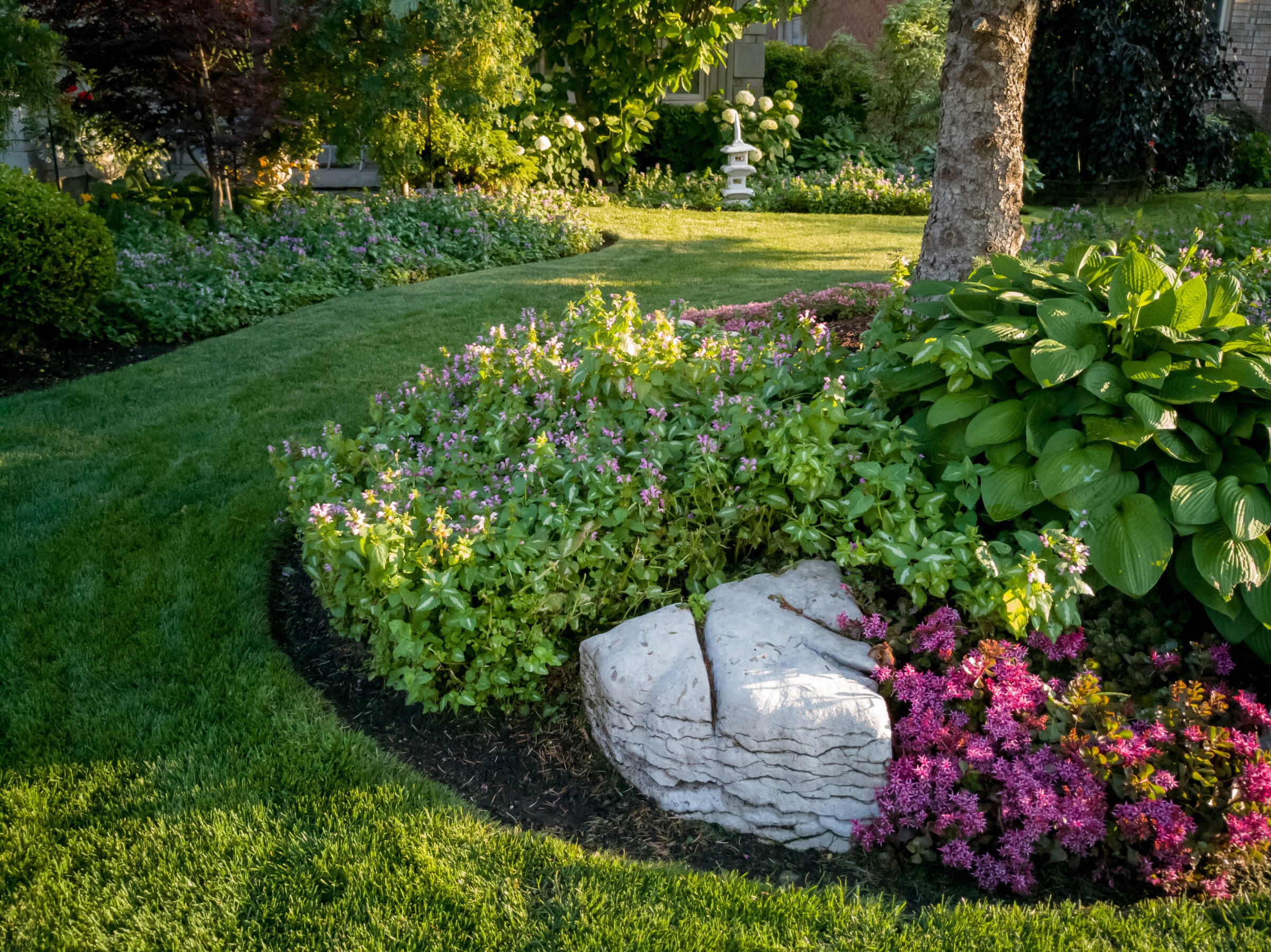 A beautifully landscaped garden features lush greenery, blooming flowers, a small stone sculpture, and well-manicured lawn surrounded by mature trees.