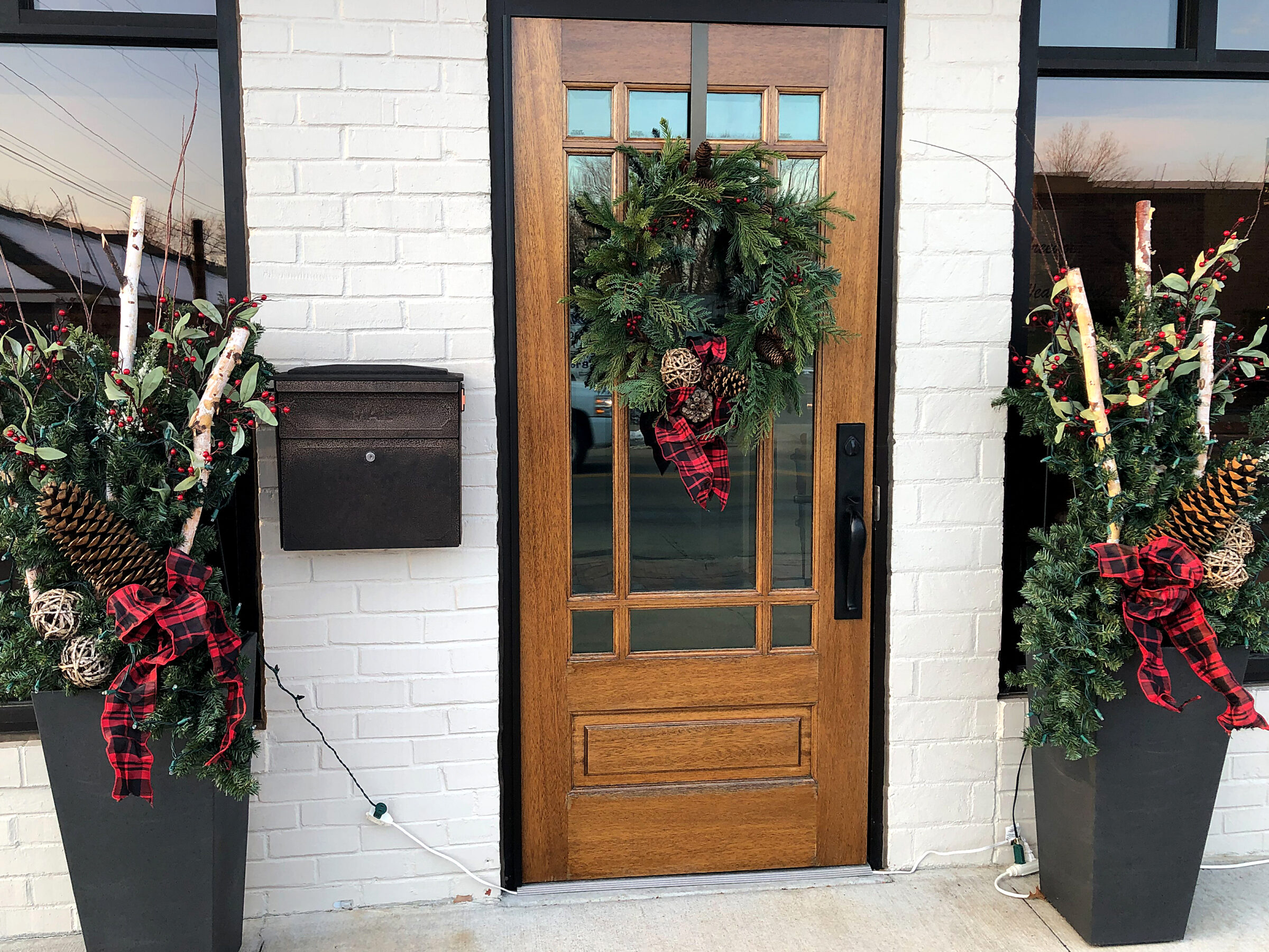 Wooden door with festive wreath, flanked by planters with holiday greenery, pinecones, and red plaid ribbons against a white brick wall.