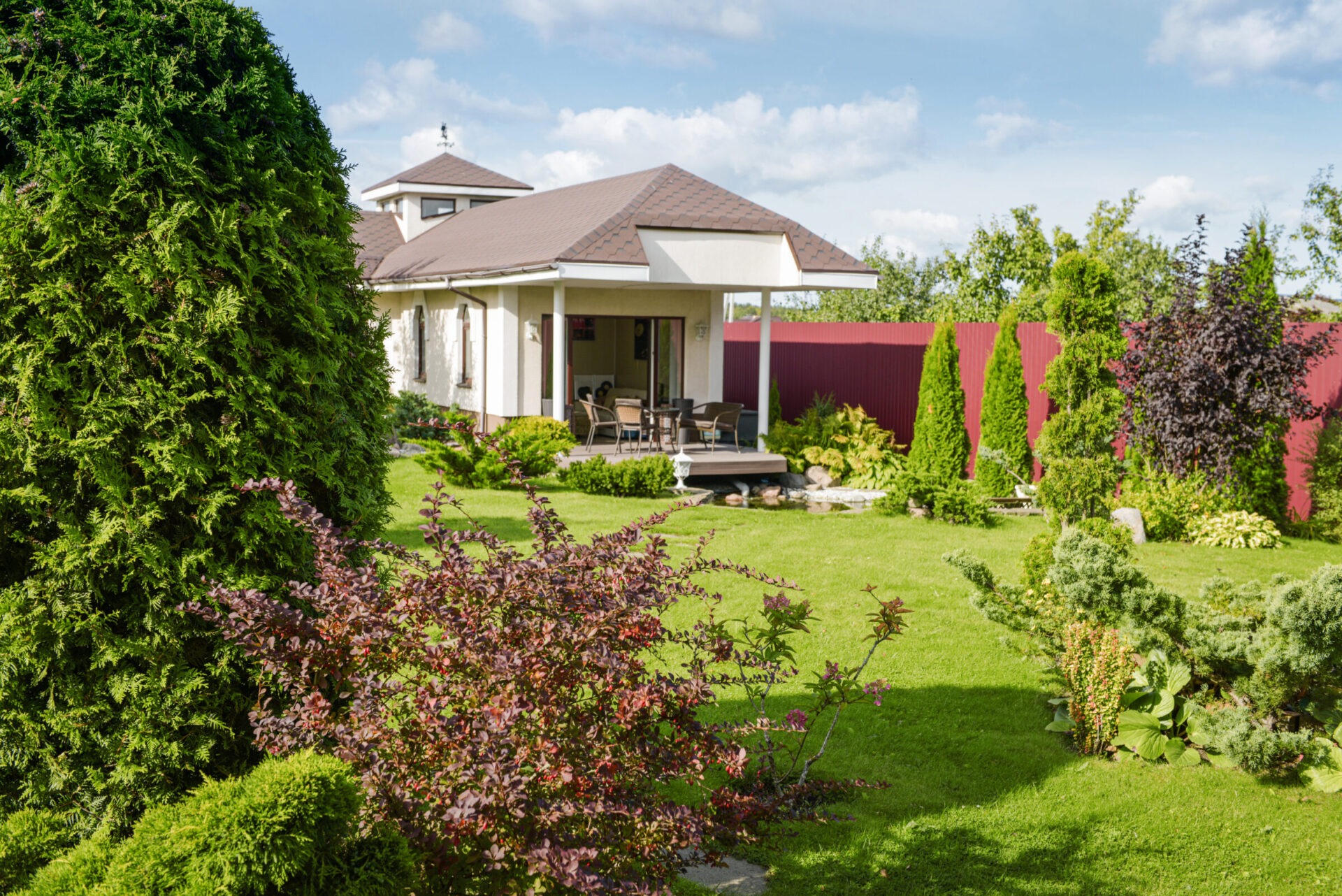 A small back yard on a sunny day with many well-manicured plants, a small deck with chairs, and a pond.