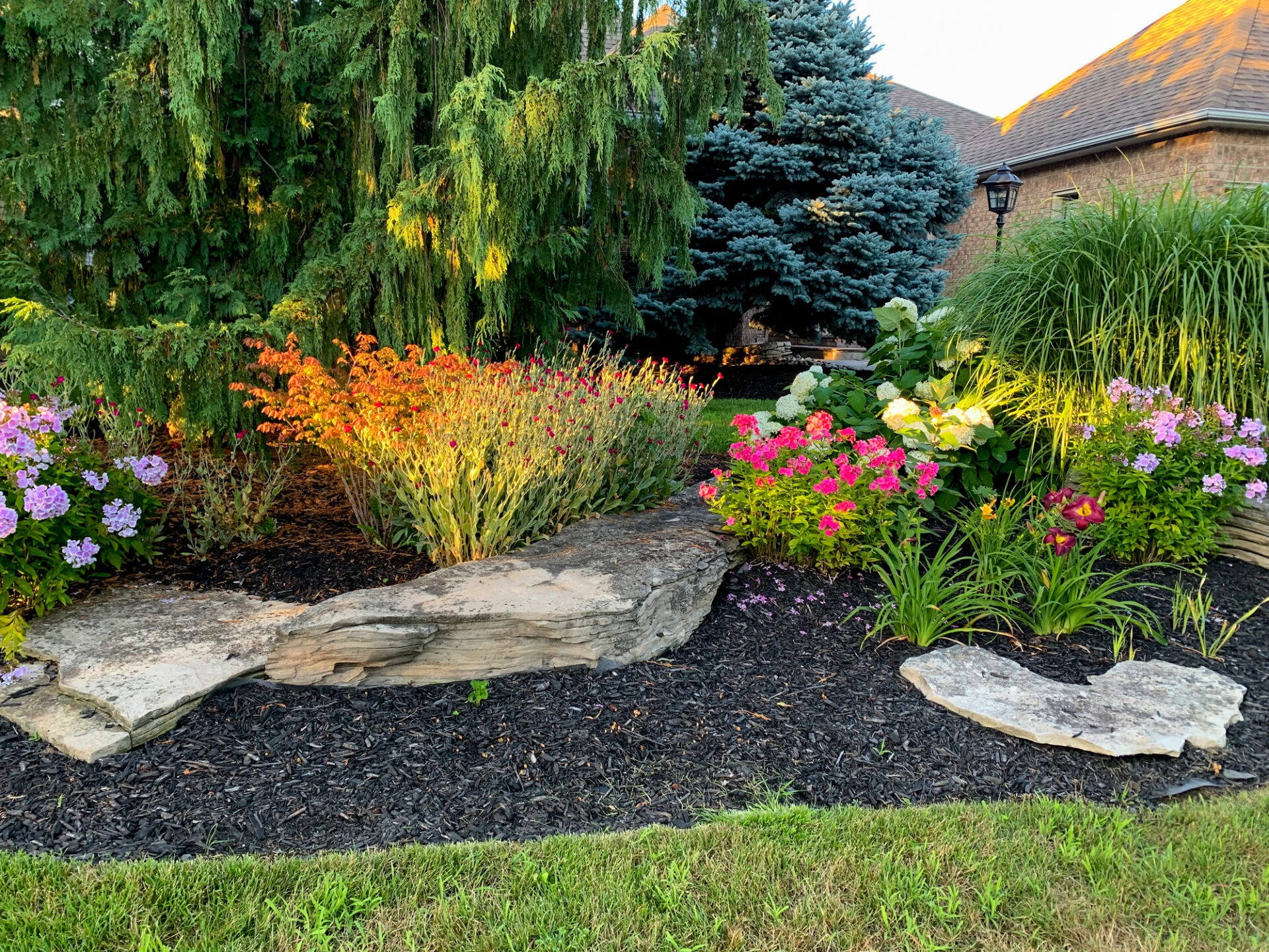 A vibrant garden scene with colorful flowers, a variety of evergreens, stone edging, and a lamp post near a brick building.