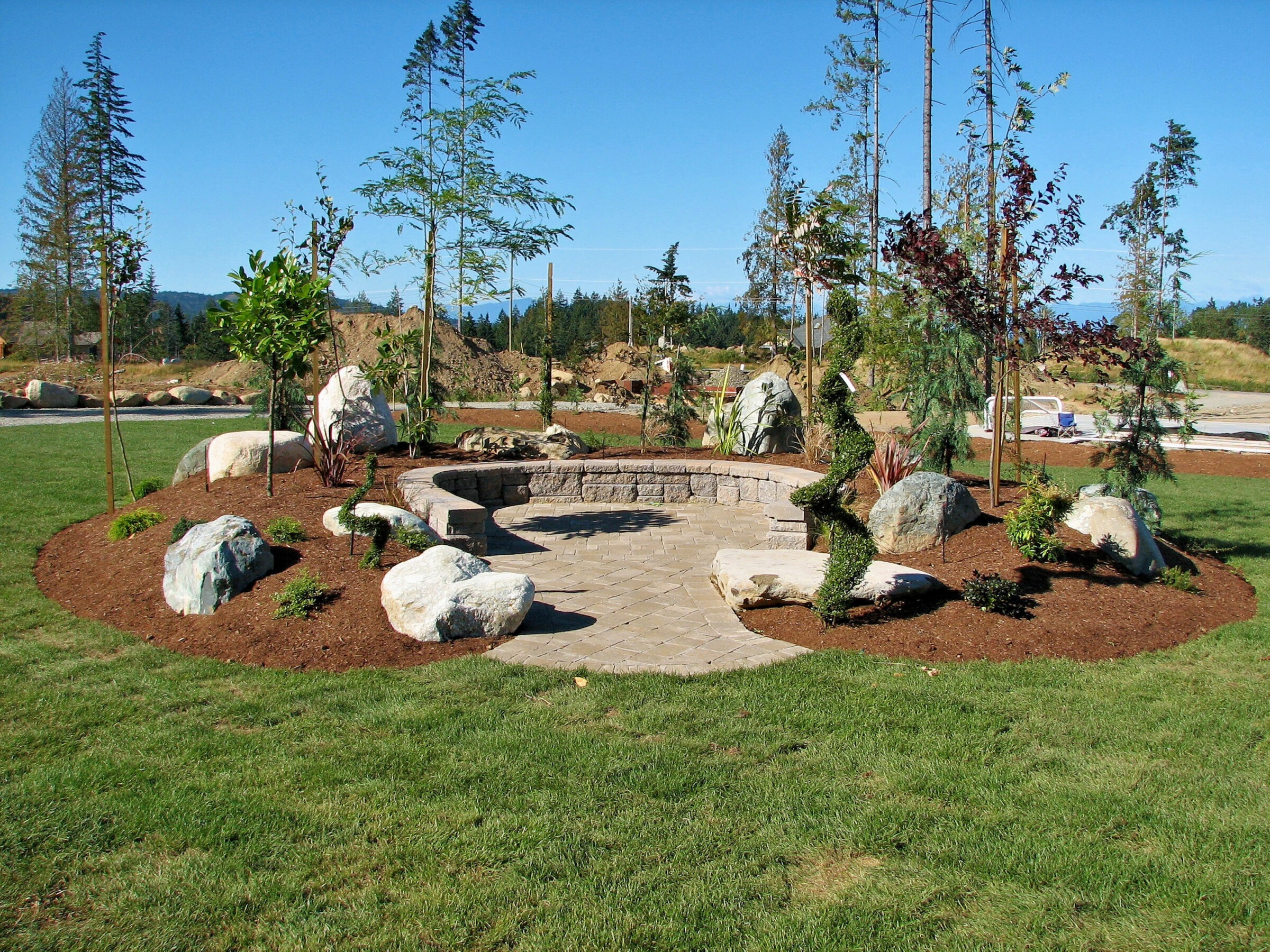 A landscaped garden area with stone pathway, surrounded by rocks, young trees, and well-manicured shrubs under a clear blue sky.