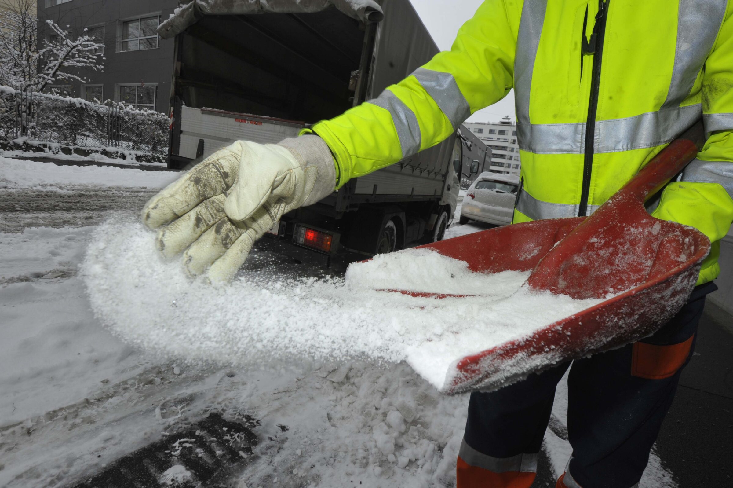 A person in a yellow jacket spreads salt with a shovel on a snowy road, near vehicles and a building, to prevent ice.